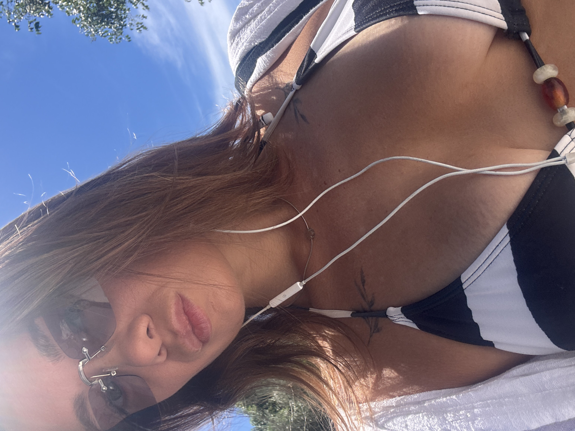 A woman is taking a selfie outdoors in Kamenovo, Montenegro. She is wearing a black and white striped bikini, a white cover-up draped over her shoulders, and sunglasses. White earbuds are in her ears and draped over her chest. She has a small tattoo on her upper chest. The background shows a clear blue sky with some tree branches visible on the right side. The lighting suggests it is a sunny day.