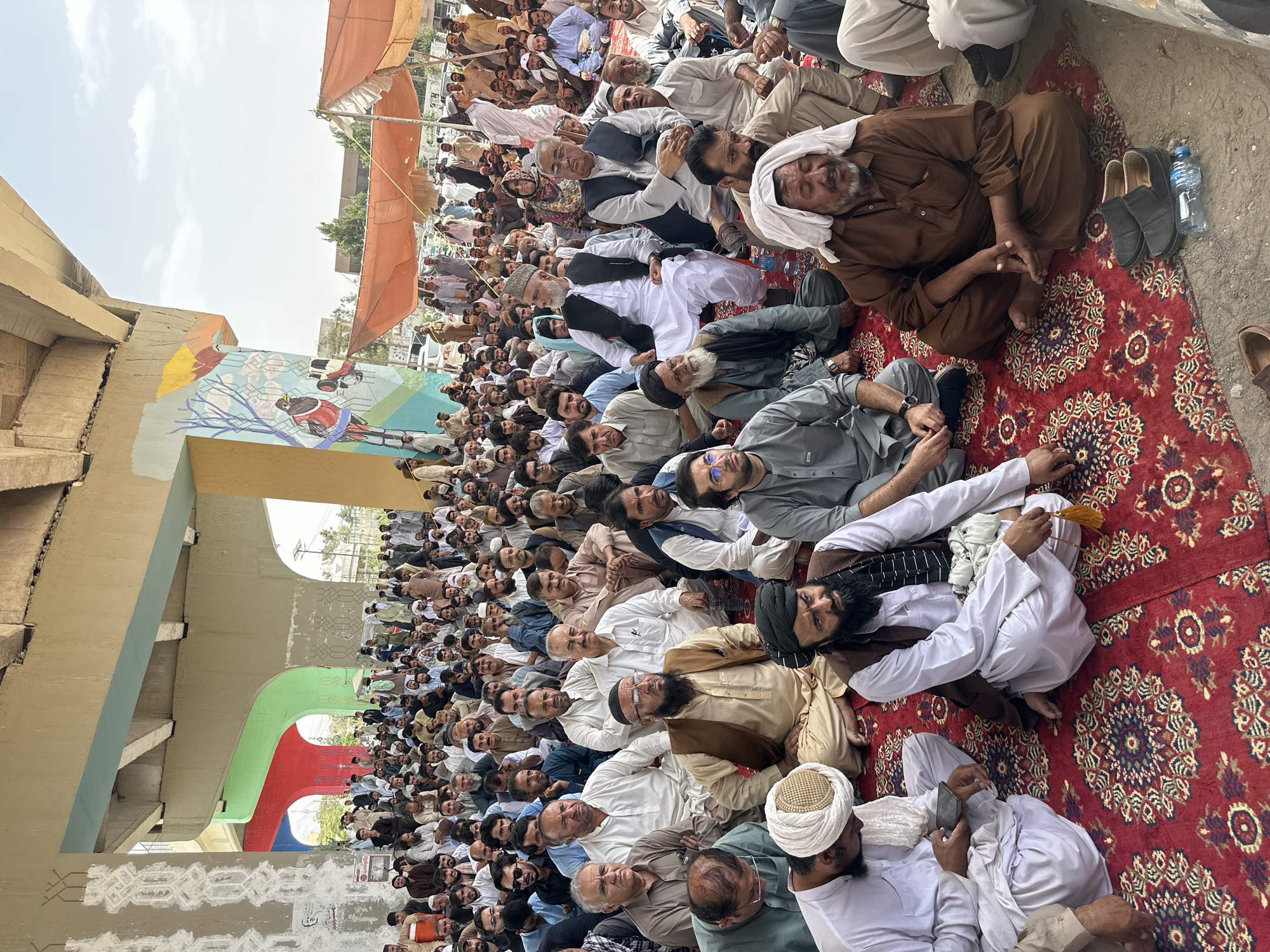 A large gathering of men are seated on a red carpet under a bridge in Quetta City Tehsil, Pakistan. The men, dressed in traditional clothing, are mostly older and appear to be participating in a religious or political event. The bridge pillars are painted with colorful murals. In the background, tents are set up, and there are parked vehicles. The scene suggests a public gathering, possibly a protest or a religious congregation.