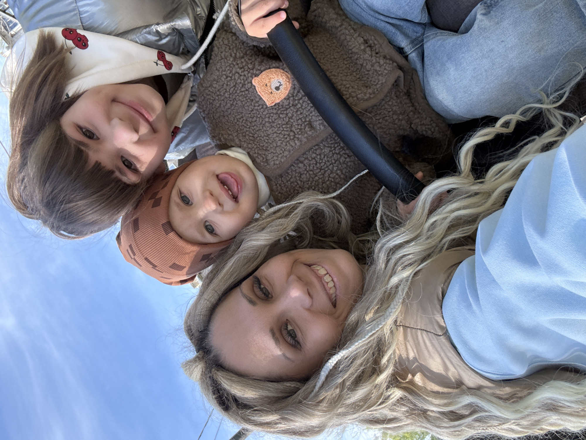 The image shows three individuals: two females and one child. One female has shoulder-length brown hair, wearing a silver puffer jacket with a hoodie featuring cherry patterns. The child, presumably younger, is wearing a brown hat and a brown, fleece-like jacket with a bear patch. The other female has long blonde wavy hair. They appear to be on some sort of ride, as a black bar is visible. The background shows a clear blue sky. The specific location of Rusanovo, Russia, cannot be definitively confirmed based solely on the image.