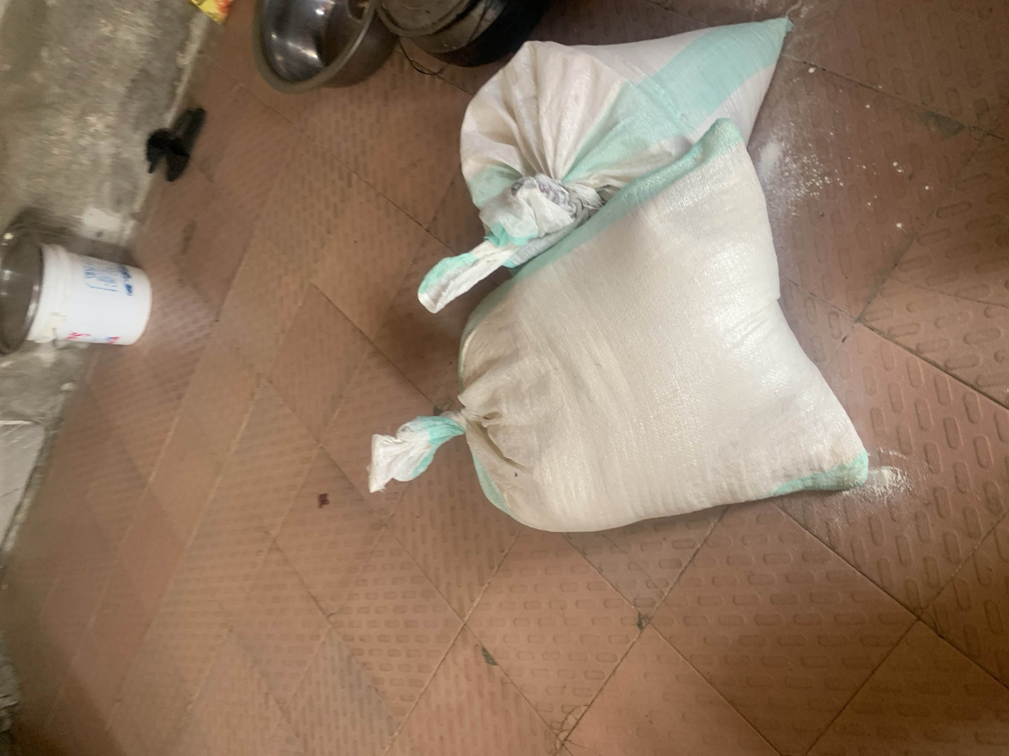 The image depicts an indoor scene with two large white sacks of what appears to be rice or flour, tied at the top with green and white fabric. The sacks are resting on a reddish-brown tiled floor. To the left of the sacks, a white plastic bucket with a lid is visible, and to the right, a metal bowl is present. The floor tiles have a patterned texture. In the background, there are plain concrete walls and some dark-colored footwear. The lighting suggests an interior space. The scene is likely in a home or storage area in Jauro Yinu, Nigeria, as indicated.