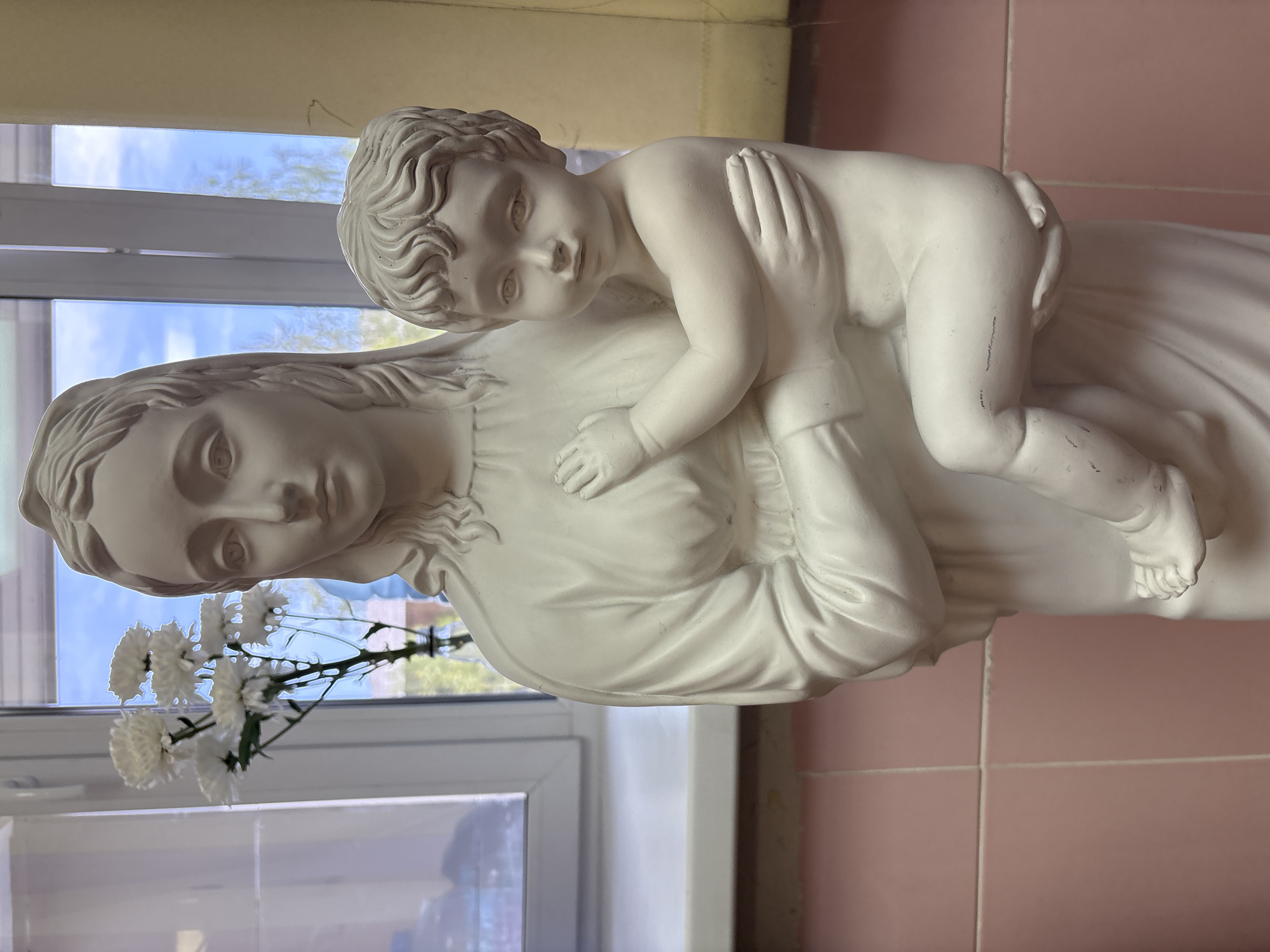 A medium, eye-level shot shows a white statue of a woman and child, likely Mary and Jesus, located in Unknown, Unknown. The statue is near a window with white trim. A small bouquet of white flowers sits in front of the statue near the window. The background to the right of the statue features pink tiled walls. The sky, visible through the window, is blue with light clouds.