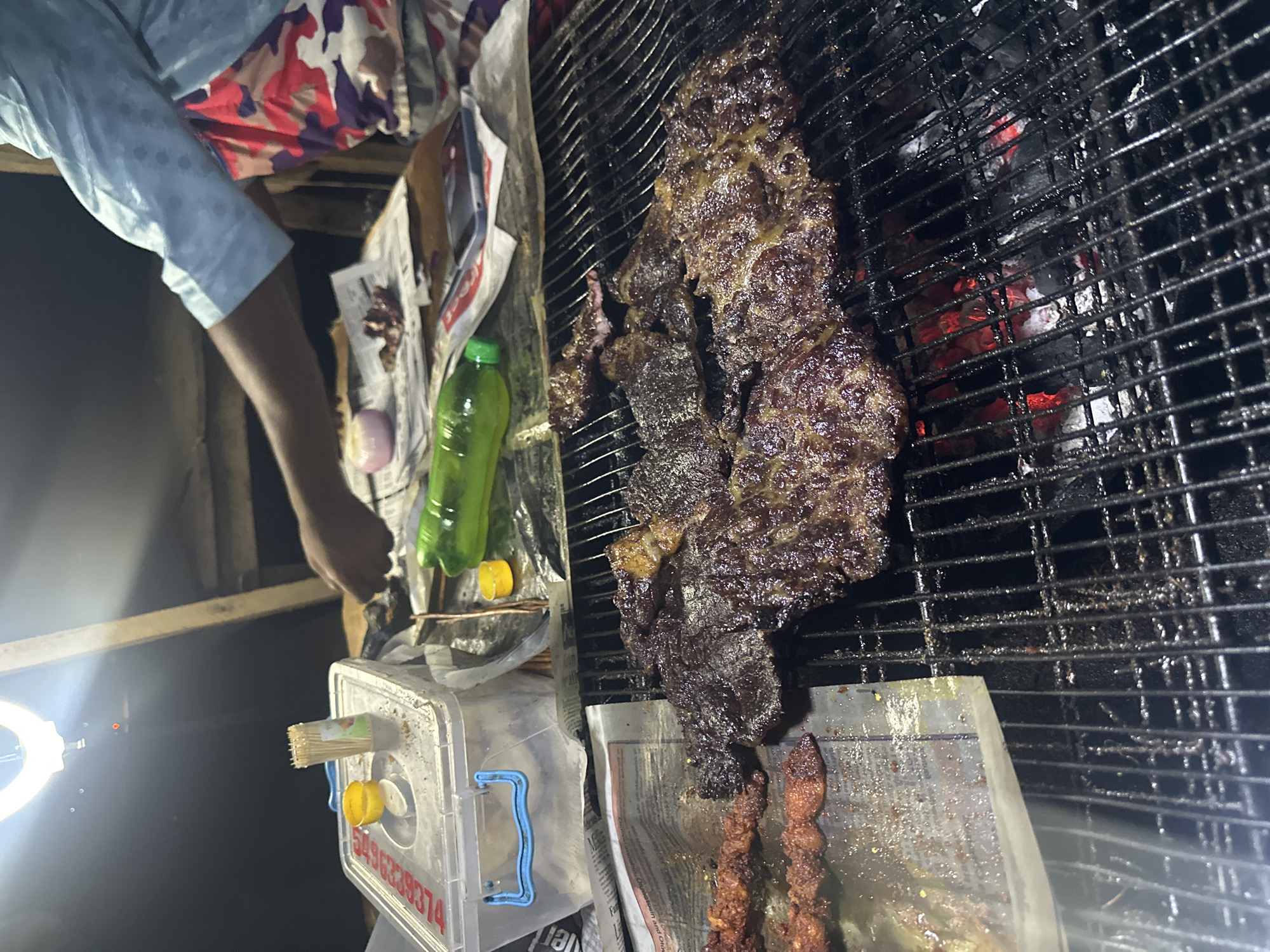 A nighttime street scene in Abuja, Nigeria features a man tending to a grill. The grill, made of black metal bars, has glowing red embers beneath. Large pieces of grilled meat are laid across the grill, and skewers with smaller pieces of meat are visible at the bottom. The man, wearing a light blue shirt and patterned shorts, is near the grill. An onion and a green bottle are nearby. A transparent container labeled with a phone number sits next to the grill. A ring light is positioned to illuminate the scene. Newspaper lines surfaces.