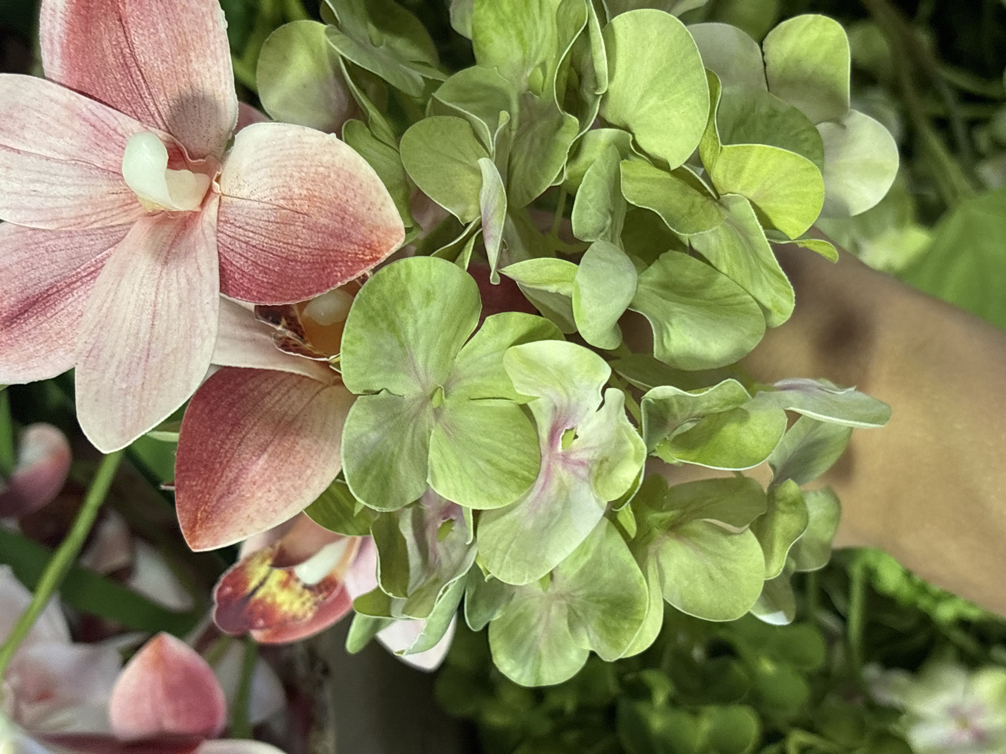 A close-up shot of artificial flowers fills the frame. A pink orchid is positioned on the left, and a cluster of green hydrangea-like flowers occupies the center and right portion. A human hand is partially visible behind the flowers. The specific location in Perm, Russia, is not discernible.