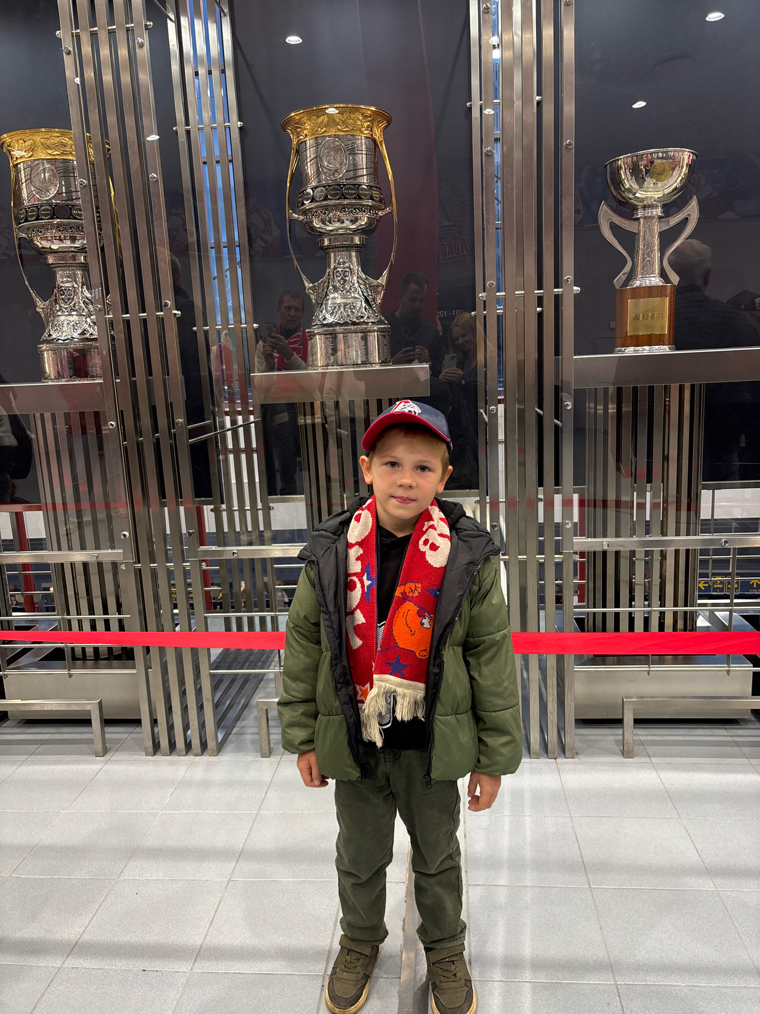 A young Caucasian boy with light brown hair, wearing a green puffer jacket, dark pants, a red and white scarf, and a red baseball cap, stands in front of a display of trophies. He is facing the camera with a neutral expression. Behind him, three large, ornate trophies are displayed in glass cases with metal supports. Reflections in the glass show other people and trophies. The environment appears to be indoors, with a tiled floor and a red barrier in the foreground. The location is identified as a trophy hall in Rusanovo, Russia.