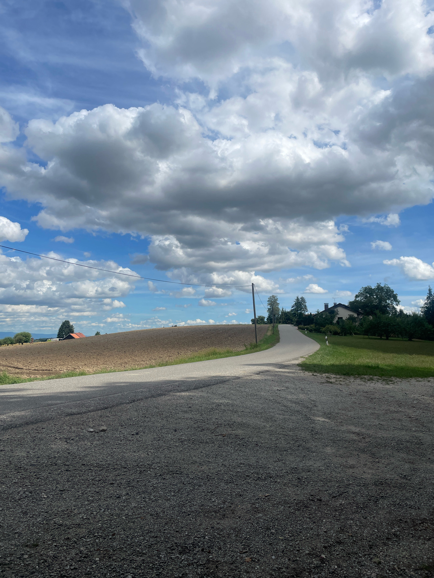 The image depicts a rural landscape with a paved road curving through rolling hills under a partly cloudy sky. The road is the focal point in the foreground, composed of dark gravel. To the left of the road, a large, tilled brown field stretches towards the horizon. In the distance, a red-roofed building is visible amidst trees. To the right of the road, a manicured green lawn leads up to a white-colored house with dark windows, surrounded by lush trees and foliage. A utility pole stands to the left of the road further up the incline. The sky is a vibrant blue, punctuated by large, white and gray cumulus clouds. The overall scene conveys a sense of peaceful countryside.
