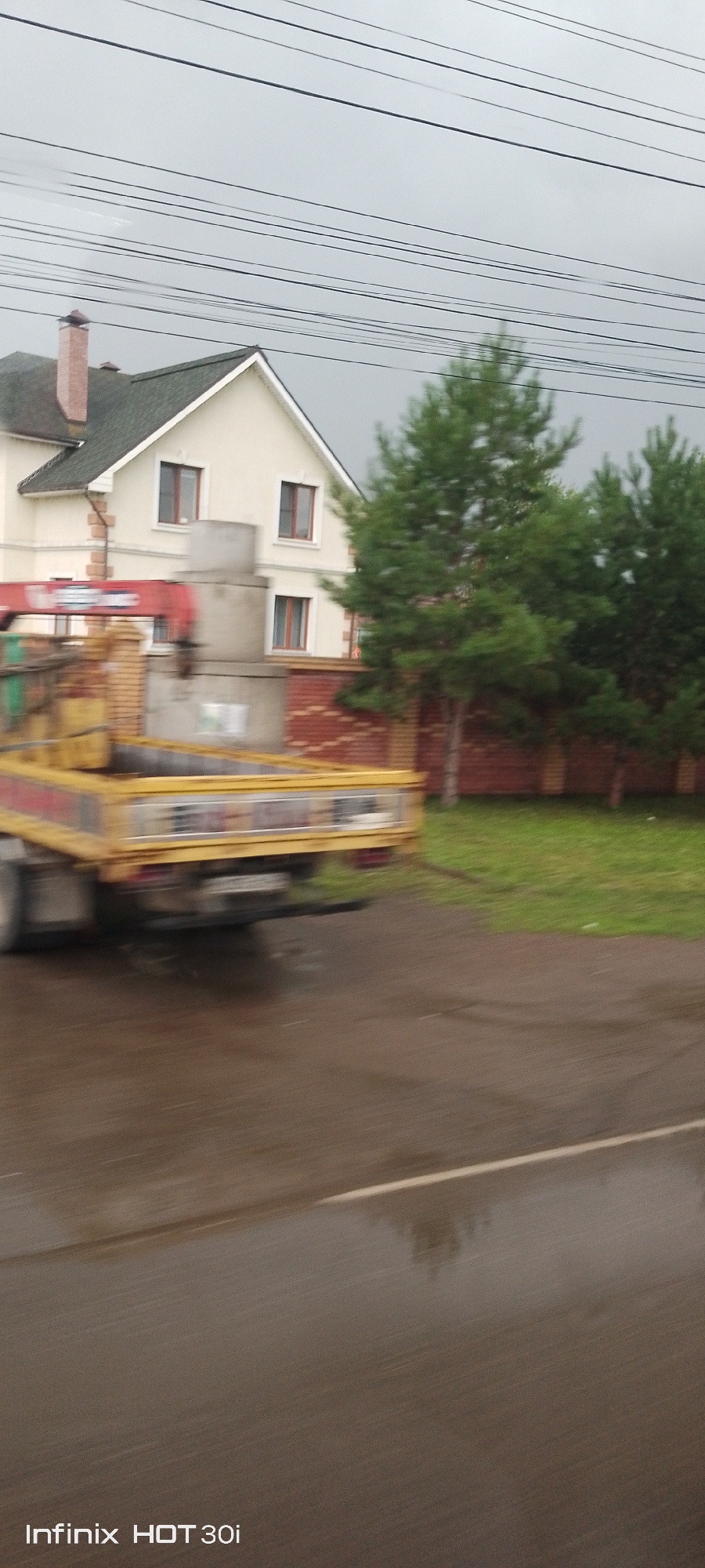 This is a photograph of a truck carrying concrete rings, moving along a wet asphalt road. The truck is yellow and has a red crane attached. In the background, there is a house with a green roof and a red brick fence. There are also pine trees and power lines. The image appears to have been taken from a moving vehicle, as indicated by the motion blur. The timestamp "Infinix HOT 30i" is visible in the bottom left corner. The scene is located in Yemelyanovo, Russia.
