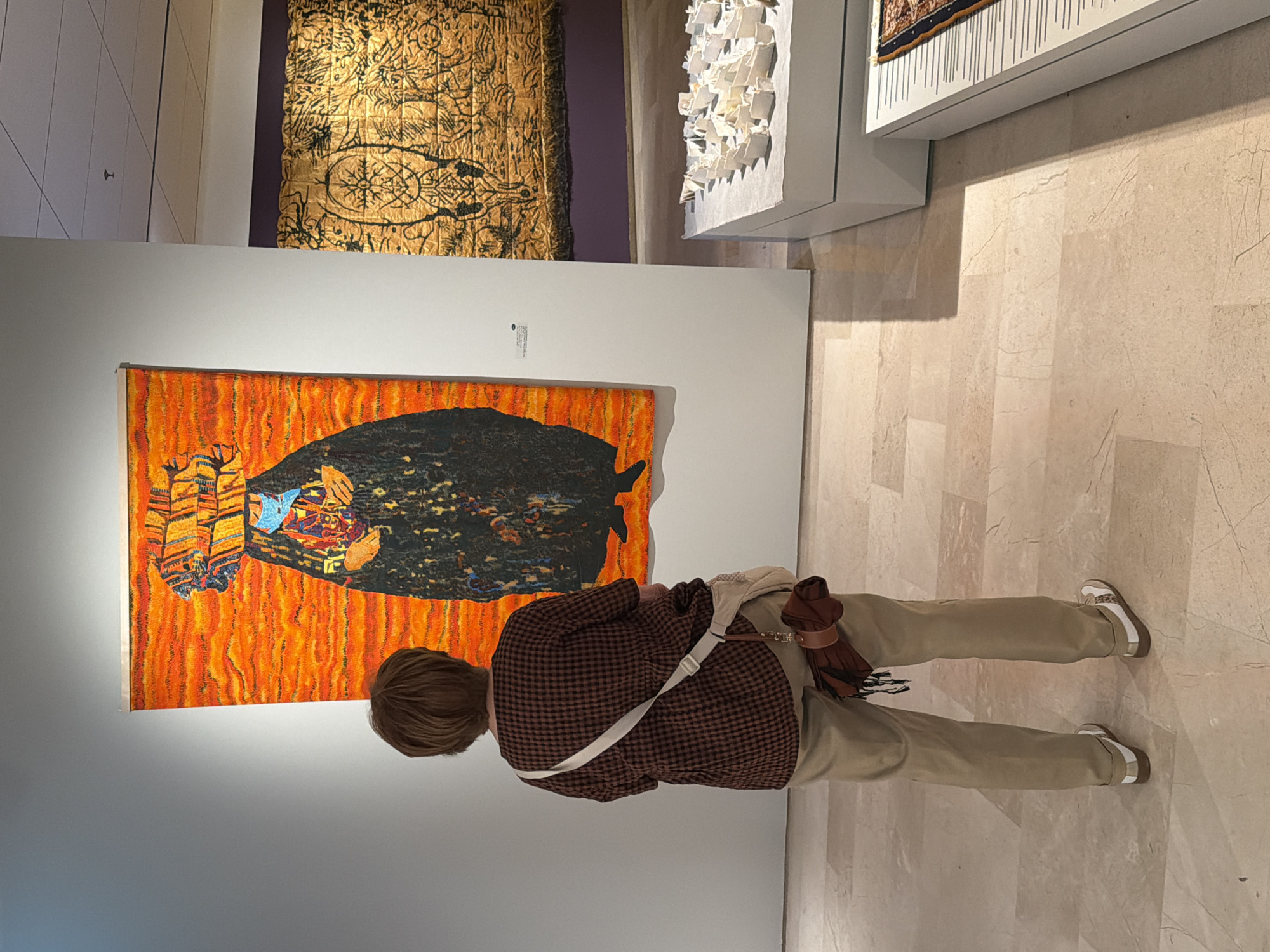 A middle-aged woman stands in an art gallery, observing an orange and black painting. She is wearing a brown checkered jacket, khaki pants, and white sneakers. She has a tan purse with a white strap and a brown accessory attached. The gallery has tile flooring, and there are several other art pieces in the background. Based on available information, there is no information of the location of the art gallery in the city of Tel-Aviv, Israel.