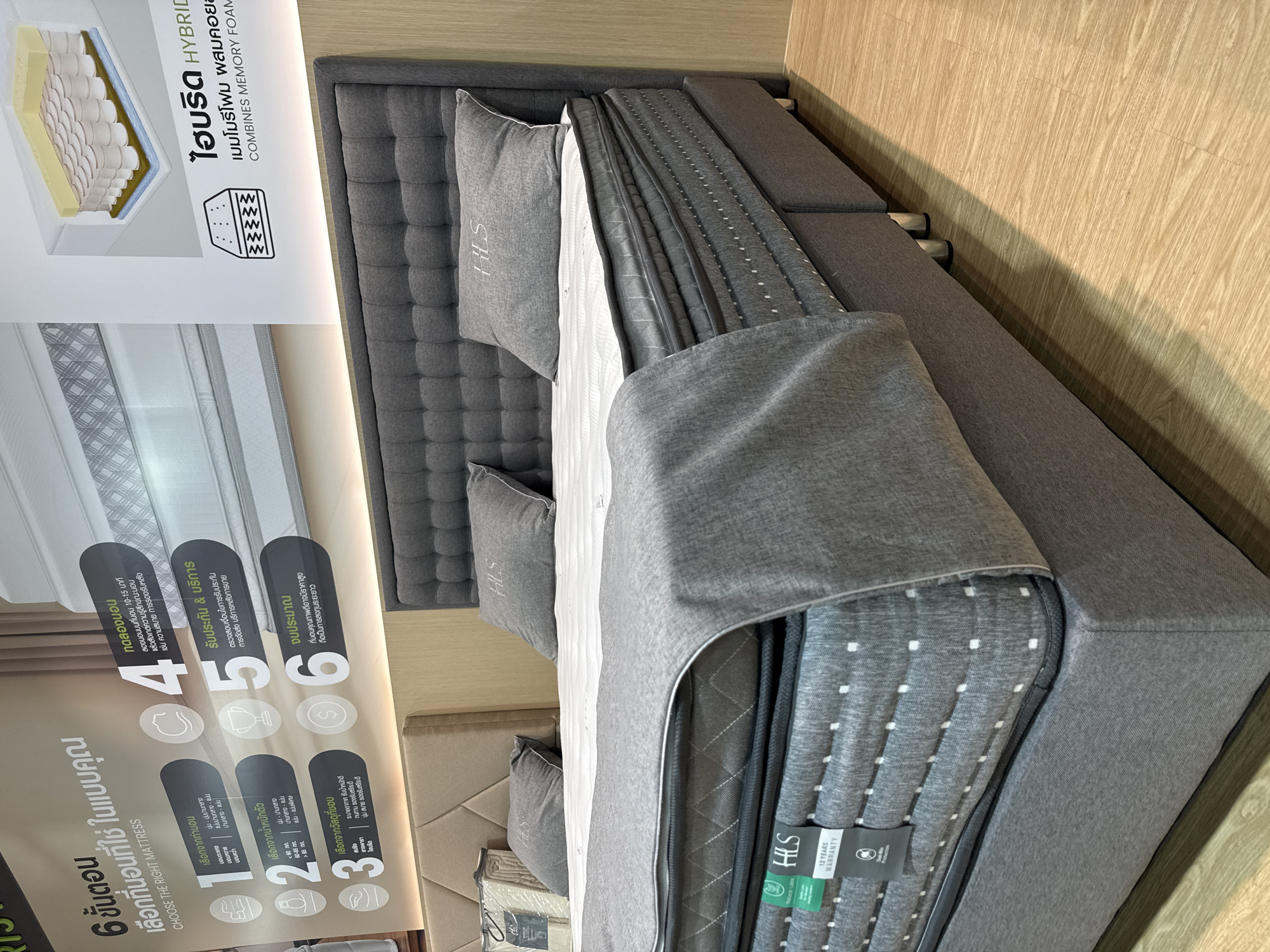 The image shows a mattress in a showroom. The mattress is gray with white stitching and is part of a bed with a gray headboard and base. There are two pillows on the bed, also gray with an embroidered logo. A gray blanket is draped over the mattress. In the background, there is a wall with a diagram of a mattress and text in Thai and English. There is also a portion of another bed visible on the left. The flooring is light-colored wood. The scene appears to be an indoor retail environment. The location is stated to be Baan Chaweng Noi, Thailand.