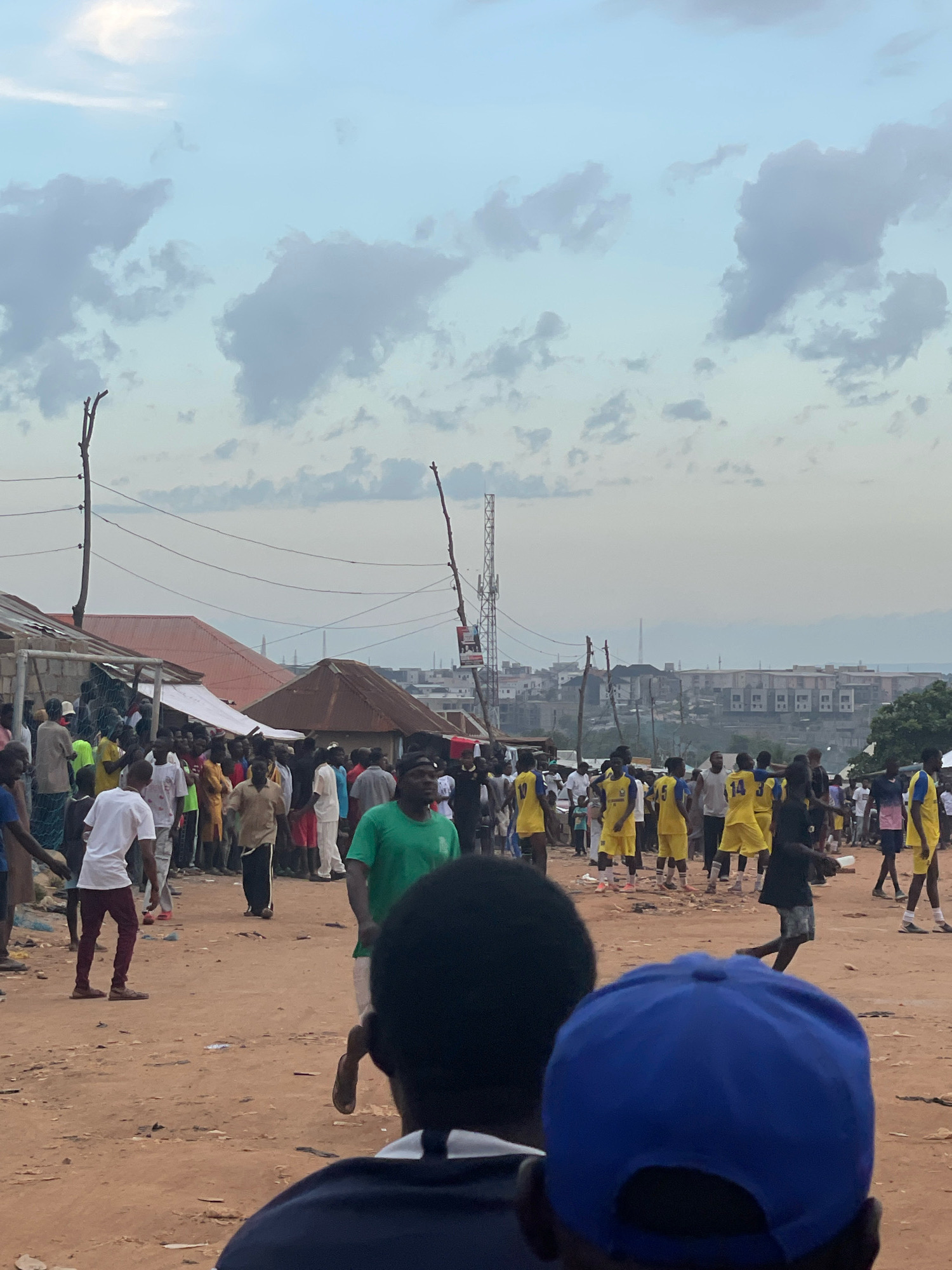 The image captures a street scene in Abuja, Nigeria, likely within a densely populated or informal settlement area. The foreground is dominated by the back of two individuals, one wearing a blue baseball cap and another with dark hair. The middle ground reveals a crowd of people gathered on a dirt road. Several individuals are wearing yellow sports jerseys with numbers, suggesting a sports event or practice is taking place. Some spectators are also present, observing the activity. The background shows a collection of buildings, some with corrugated iron roofs, and a distant cityscape with more modern structures and a telecommunications tower. The sky is overcast with scattered clouds. The environment appears somewhat dusty and unpaved.