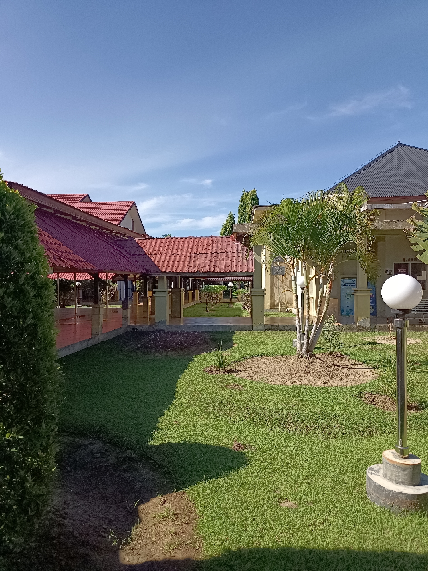 The image depicts a view of a building with a red-tiled roof and a gray metal roof, likely in a tropical or subtropical climate, as evidenced by the palm trees and lush green grass. The architecture suggests a public or institutional building, possibly a school or government office, given the signage visible on the right side of the image. The building features a covered walkway or veranda supported by pillars, with tiled flooring. A well-maintained lawn surrounds the building, with a decorative circular garden bed around a small palm tree in the foreground. A modern outdoor lamp with a white spherical light fixture stands on the right side of the image. The sky is clear blue with a few wispy clouds, indicating fair weather. The image appears to have been taken during the daytime, with sunlight casting shadows across the lawn. The location is specified as Mesjid Reuntoh, Indonesia.