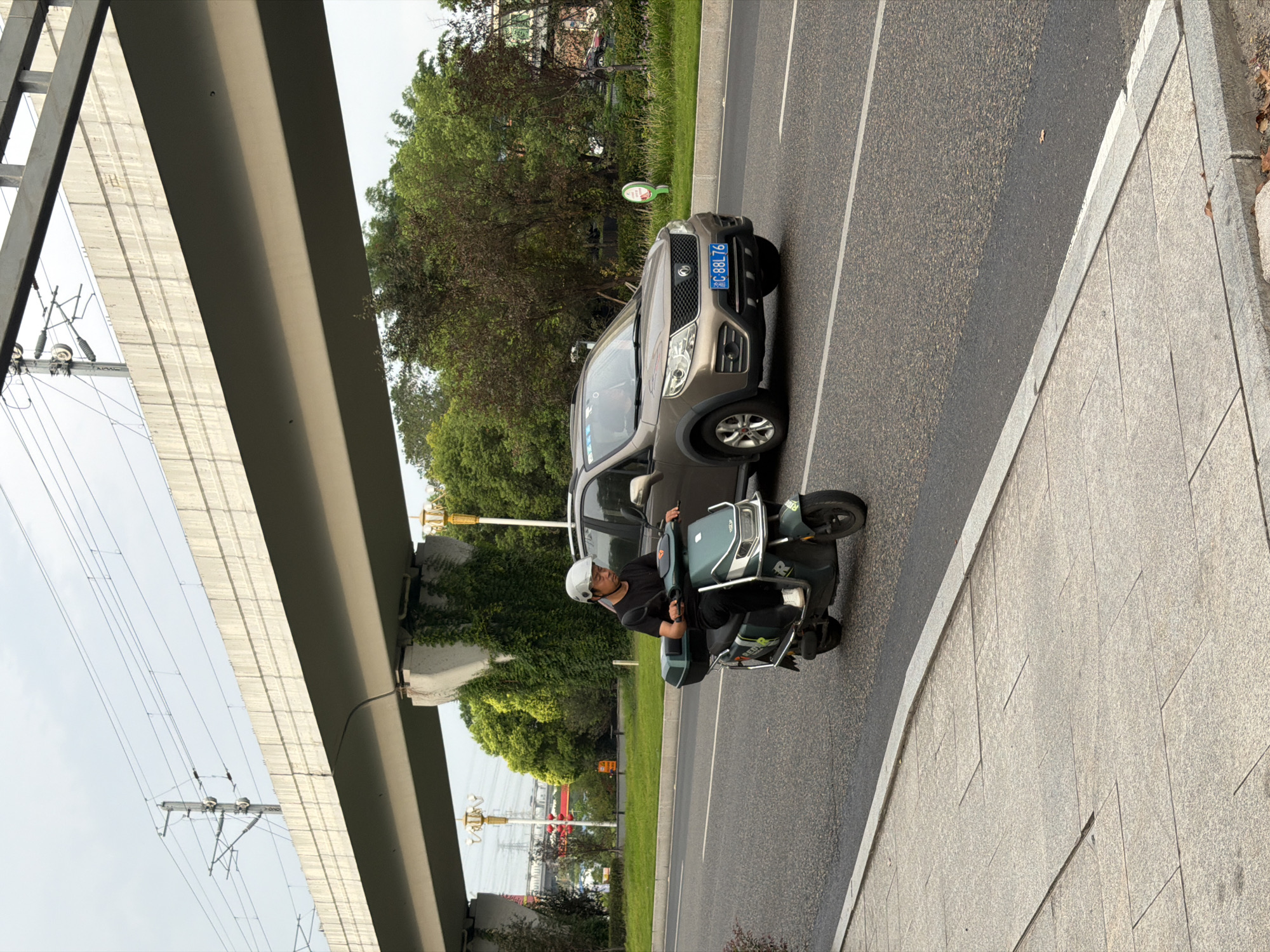In Jiangjin, China, a man wearing a black t-shirt and helmet rides a green electric scooter in the right lane of a two-lane road. He is followed by a brown SUV with a blue license plate that reads "C-88L76". The vehicles are driving under a large concrete overpass, which has train tracks and power lines above it. Green trees and foliage line the side of the road, and a sidewalk is visible in the foreground. The sky is overcast.