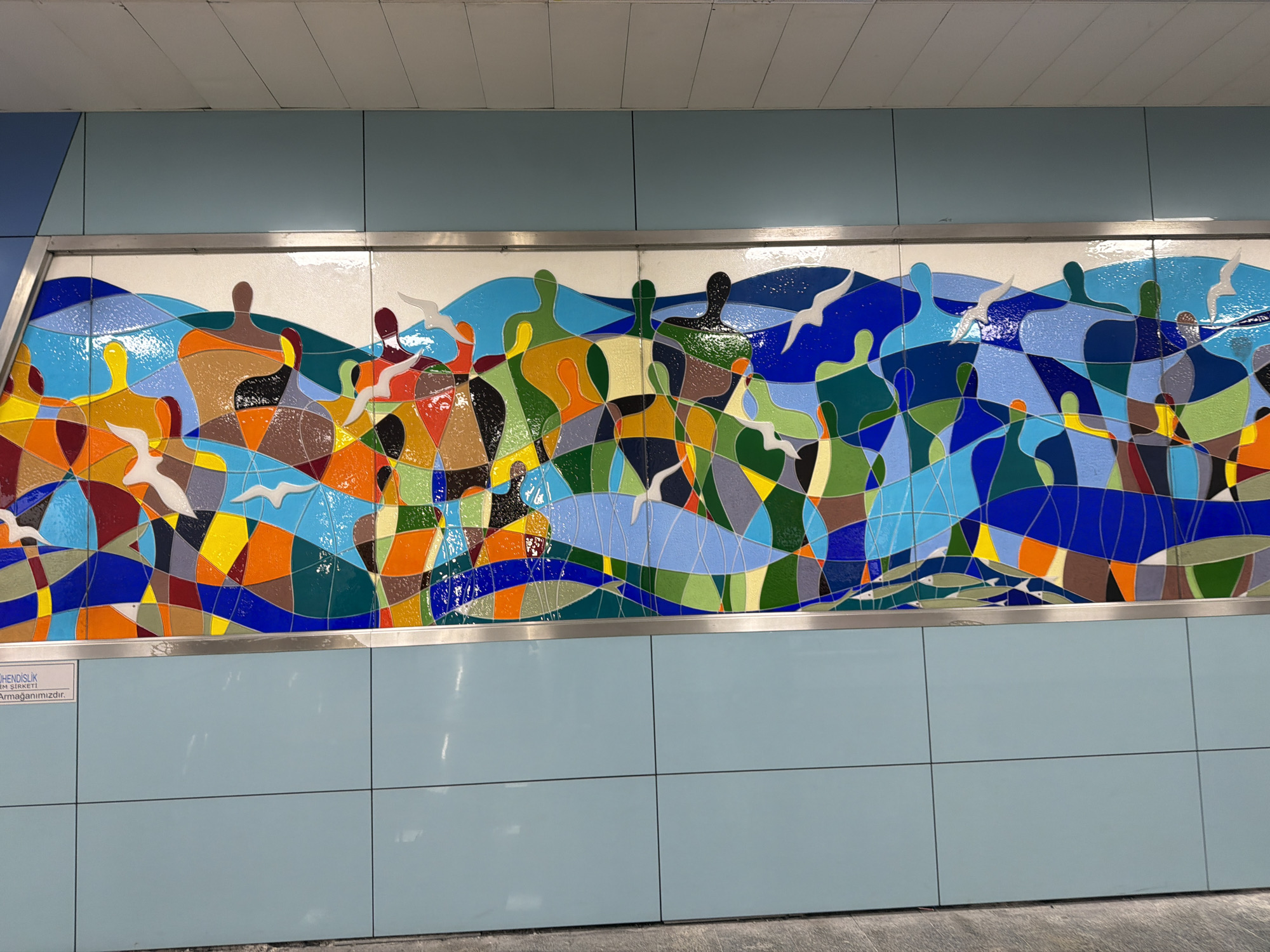 The image captures a section of an underground station in Kadıköy, Turkey. A mosaic mural, framed in silver, dominates the scene. The mural depicts stylized human figures standing on waves, interspersed with white birds, all composed of multicolored geometric shapes in shades of blue, green, yellow, orange, red, and brown. The surrounding wall is tiled in light blue, with a small sign visible on the lower left. The floor is a light grey concrete.