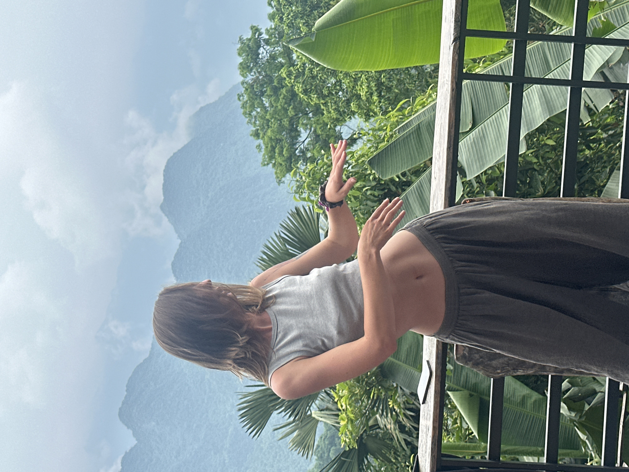 A woman with long brown hair is standing on a balcony overlooking a lush, mountainous landscape in Pù Luông Commune, Vietnam. She is wearing a grey tank top and loose, olive-green pants. Her body is angled to the right, with her arms extended outwards, as if in motion. In the background, dense green foliage and towering mountains are visible under a hazy sky. A smartphone rests on the wooden railing in front of her. The balcony is made of wood and metal.
