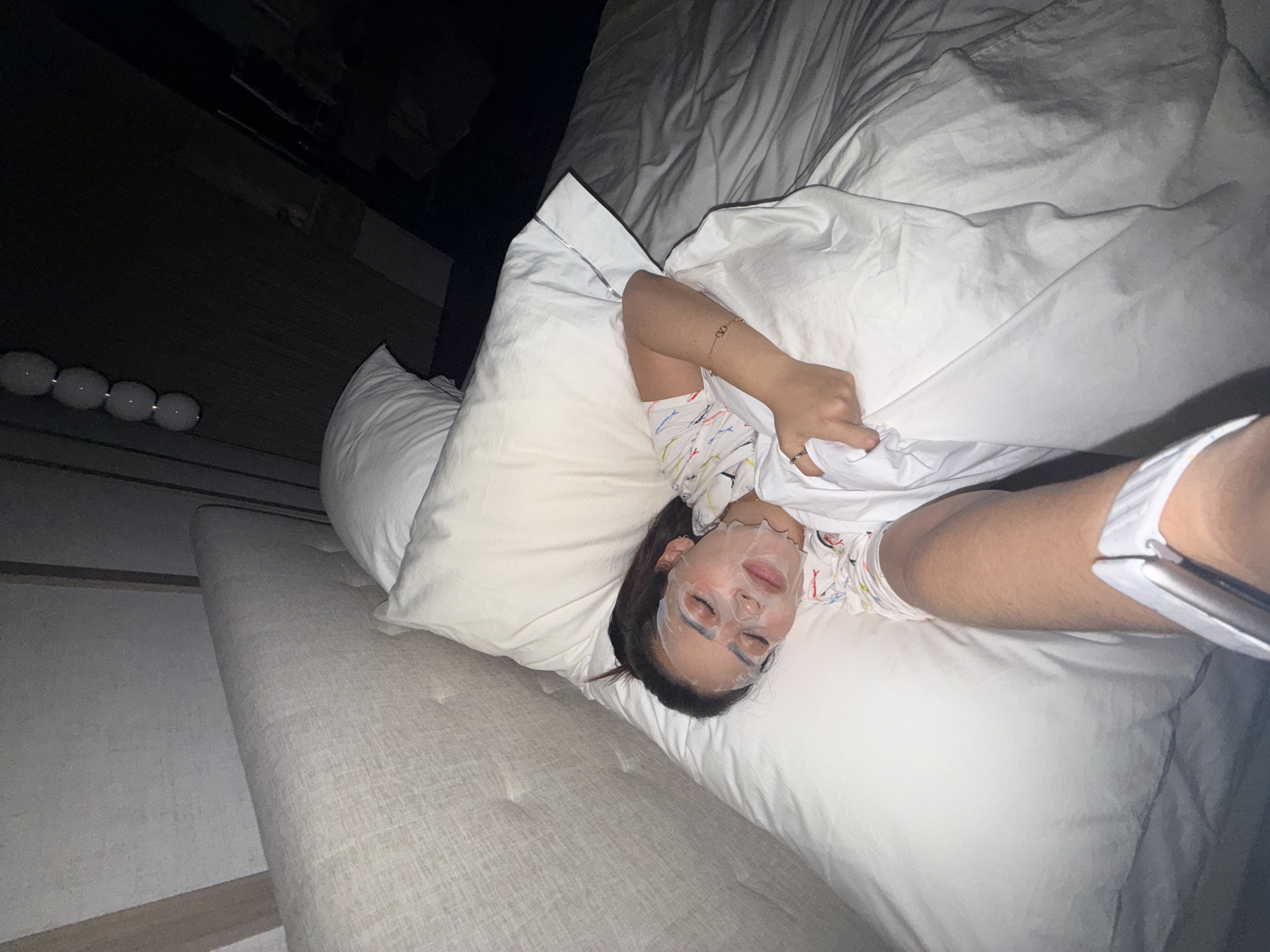 A woman with a facial mask is lying in a bed with white linens. She is holding up a white sheet. The bed has a tufted gray headboard. A string of lights is visible in the background on a dark surface. The photo was taken indoors. The location and city are not discernible from the image.