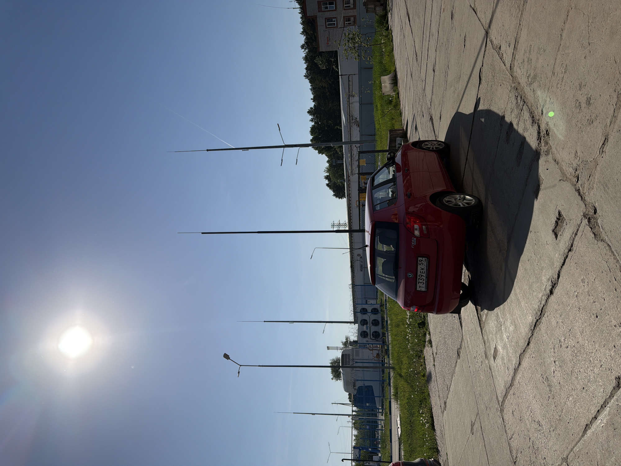 The image presents a view of an urban environment, possibly within the city of Chaikovsky, Russia. A red hatchback car is parked on a cracked concrete pavement alongside a strip of grass. In the background, there is a row of industrial-looking structures, including a series of white, container-like units and some air conditioning units. A multi-story brick building is visible in the distance. The sky is clear and blue, with the sun shining brightly, casting a shadow of the car onto the pavement. Several tall, thin poles, possibly utility poles, are seen reaching into the sky. The overall setting suggests an industrial or commercial area on a sunny day.