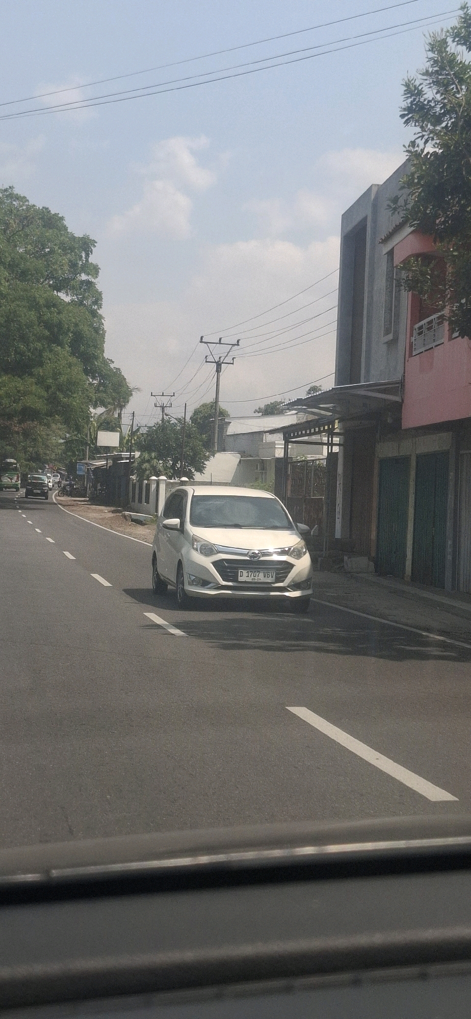 The image shows a street scene in Susukan, Indonesia. A white car with license plate D 1707 VBV is in the foreground, facing right. The car is on a paved road with white lane markings. To the right of the road is a pink and gray building with green doors. Trees and utility poles are visible in the background. The sky is blue with some clouds. The photo appears to have been taken from another vehicle.