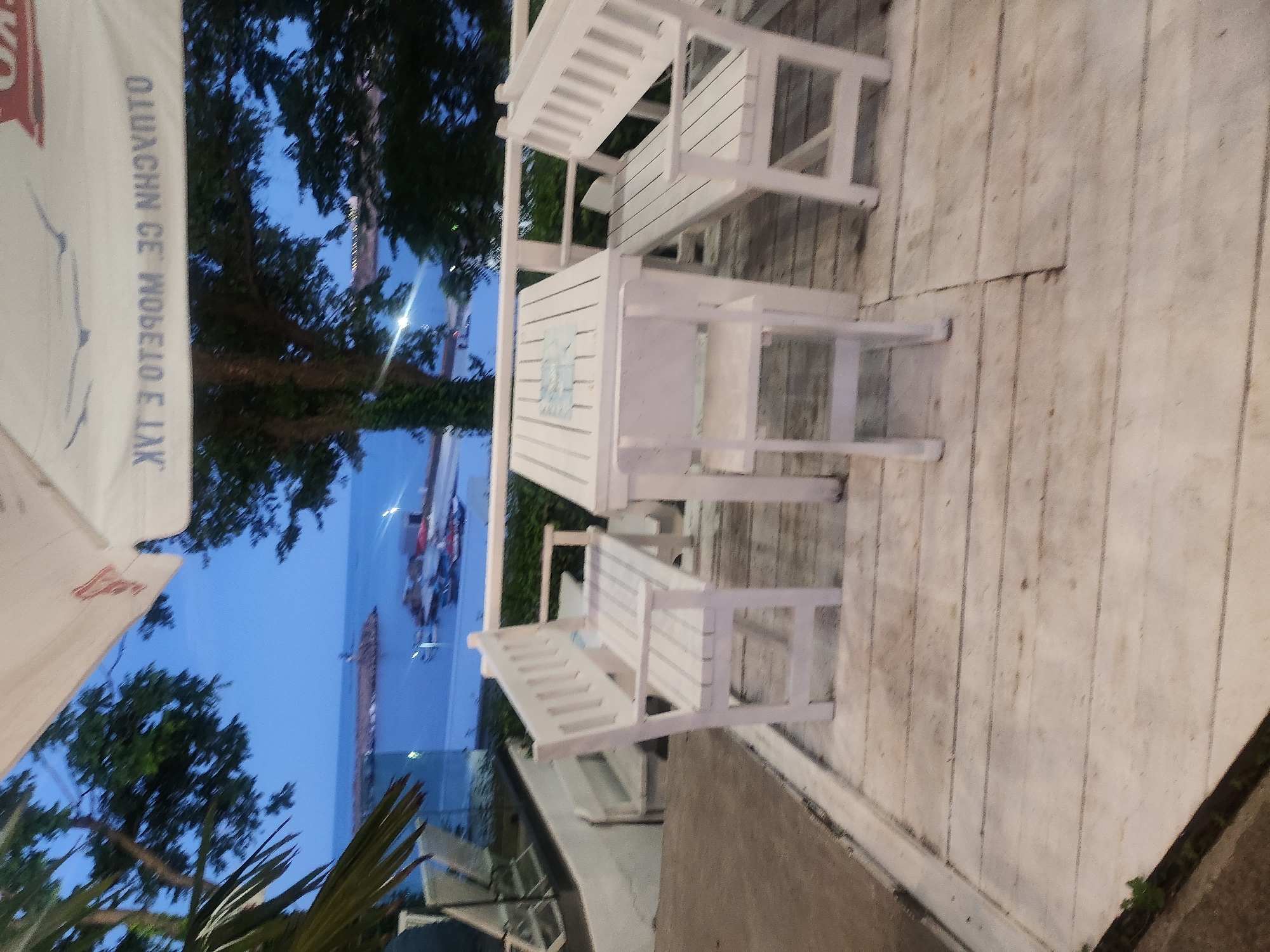 The image depicts an outdoor seating area at dusk, likely a cafe or restaurant, overlooking a body of water. The foreground features a white wooden table with two white wooden benches and a single white wooden chair. The table has a light blue decorative inlay. The seating area is on a wooden deck, with a railing visible behind the furniture. In the background, beyond the railing, is a calm body of water with several boats docked and a pier with a lighthouse-like structure. Trees line the edge of the water, and the sky is a deep twilight blue. The overall atmosphere is serene and peaceful. The scene is consistent with a location in Tsarevo, Bulgaria, known for its coastal scenery and waterfront establishments. There are no people visible in the image.