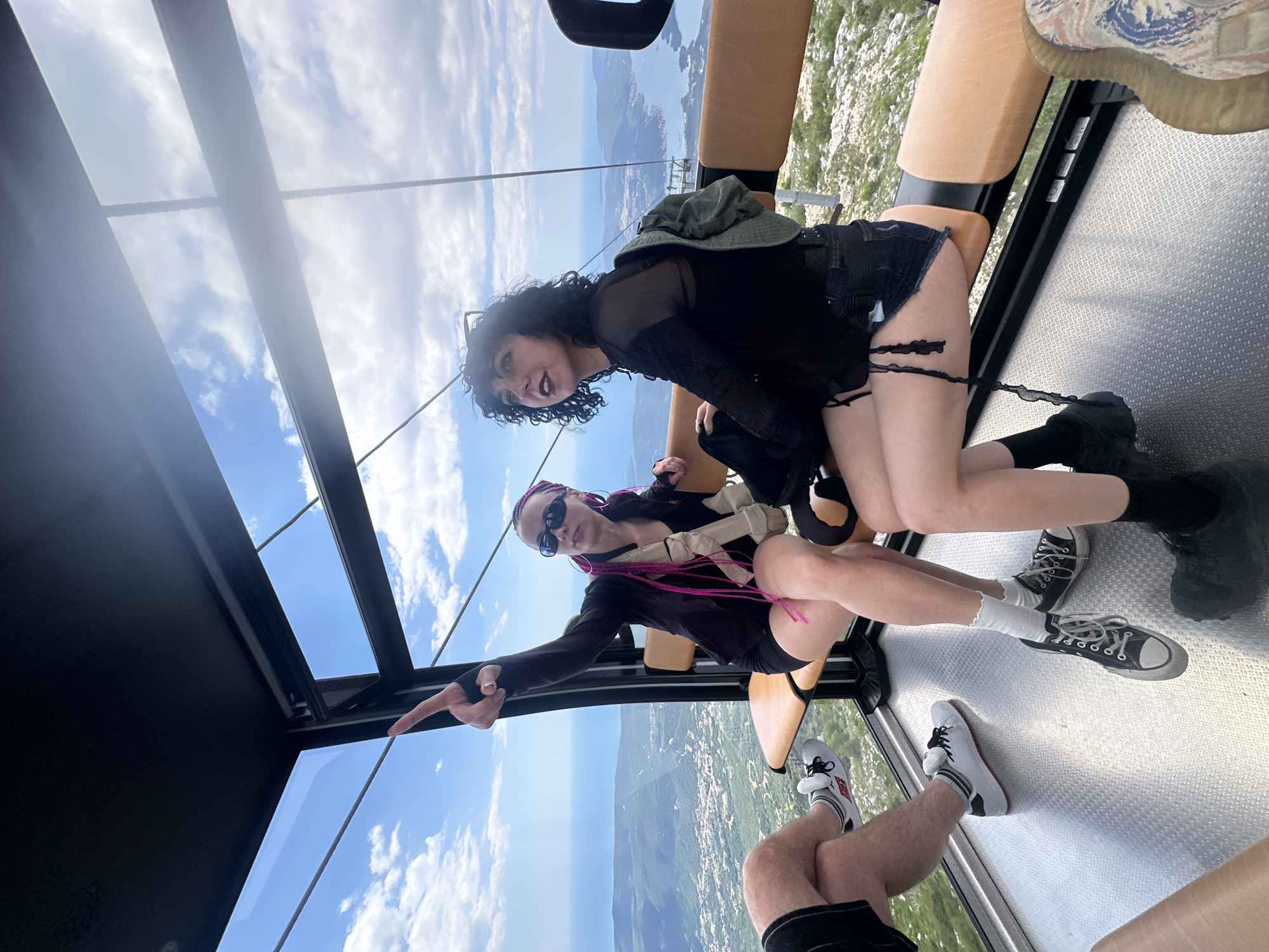 Two women are inside a cable car, possibly on a ride ascending or descending a mountain. The sky and a coastal town, potentially Škaljari, Montenegro, are visible through the cable car's windows. One woman, with curly dark hair, is wearing a black top and shorts. The other, with pink braids, has on sunglasses and is dressed in a black jacket and shorts, pointing outside. Below, the legs of another person are visible. The cable car has tan-colored seats and a metal floor.