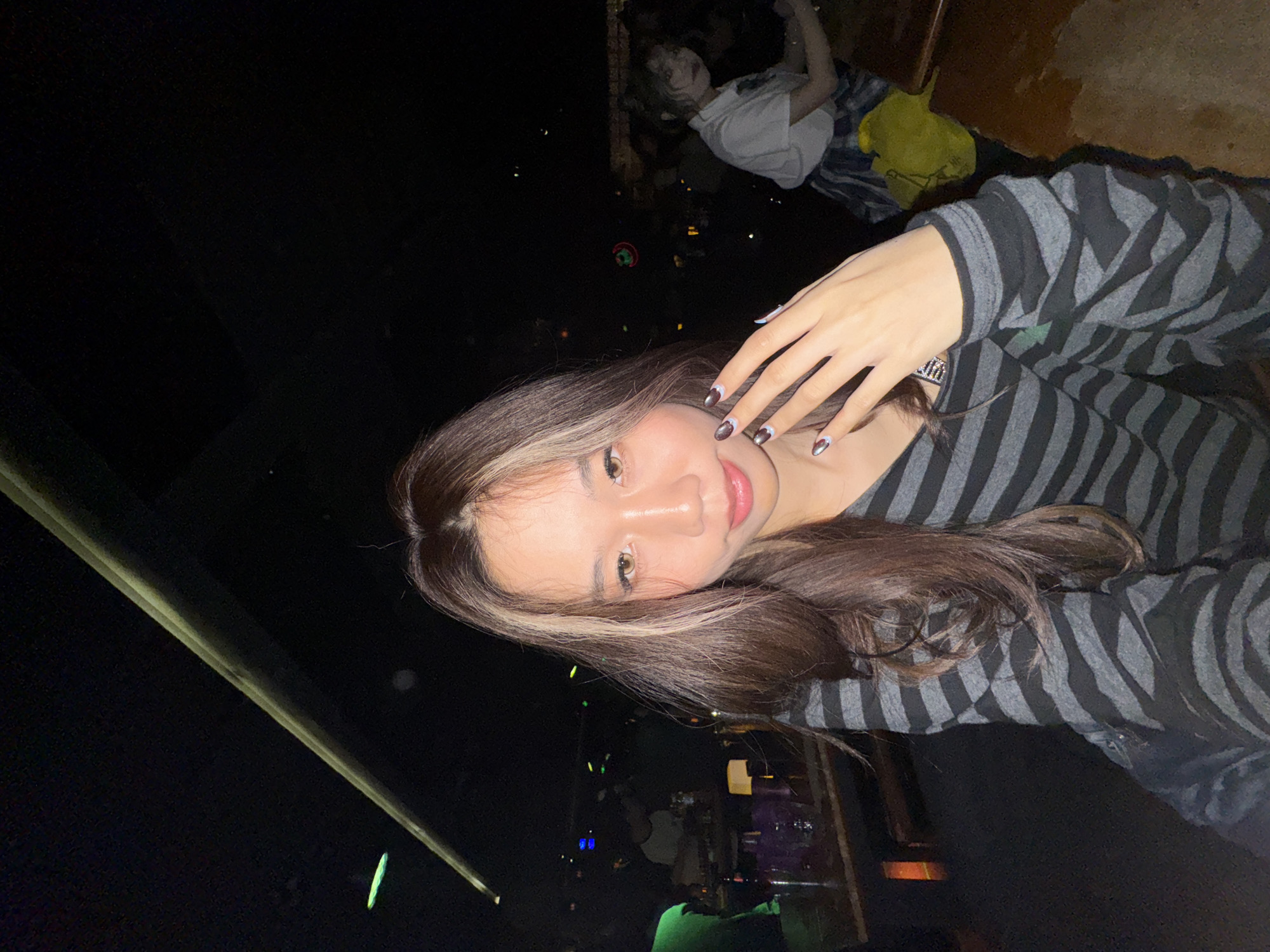 An image taken in a dimly lit indoor establishment, identified as being in Liangjiang, China, depicts a young Asian female in the foreground, facing the camera directly. She has long brown hair with prominent lighter-colored streaks framing her face and brown eyes. She is wearing a V-neck, long-sleeved top with horizontal grey and black stripes. Her left hand is raised towards her face, with fingers slightly splayed, displaying dark nail polish featuring iridescent or metallic tips. Her expression is a slight smile.
The background is dark and out of focus, suggesting a nightclub or bar environment. To the viewer's left, indistinct figures are visible, including one wearing a green shirt, situated behind a wooden table. On this table, several purple-tinted glasses or bottles are present, alongside a glowing orange rectangular object, possibly a lamp or decorative element. In the far background, various small, colored lights (blue, green, red) are discernible. To the viewer's right, a blurry female figure is seated, wearing a white short-sleeved top and a plaid skirt, with a bright yellow bag near her. A dark wooden table surface extends into the foreground on the right. A long, light-colored linear structure, possibly a ceiling beam or lighting fixture, extends diagonally across the upper portion of the frame. The scene is illuminated primarily by a direct light source on the central figure, casting the background into deeper shadow.
