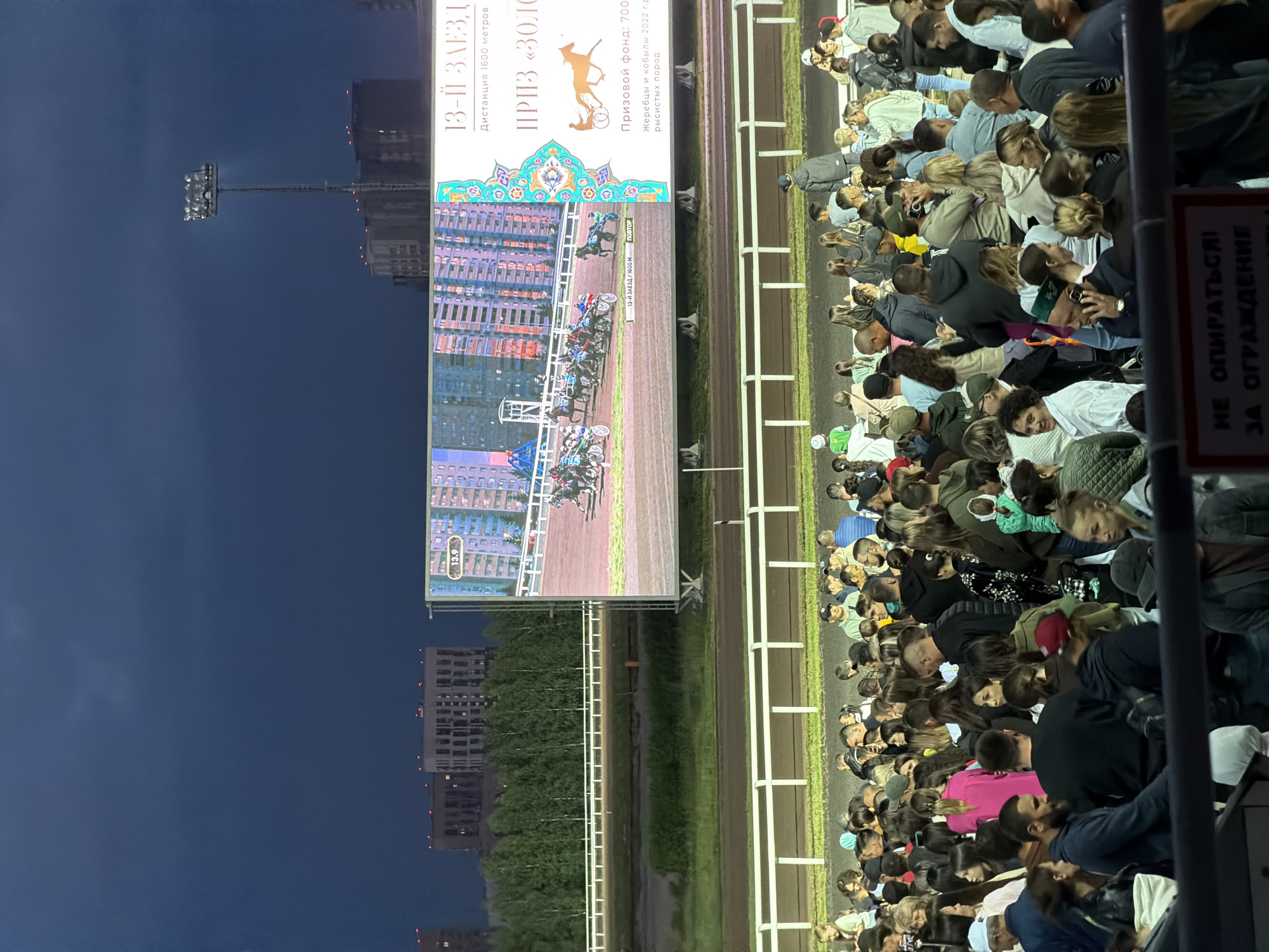 The image shows a crowded outdoor event, likely a horse race, taking place at dusk or night. A large jumbotron screen displays a horse race in progress, with several horses and sulkies on a track. Text on the screen indicates "13-й ЗАЕЗД" (13th race), "Дистанция 1600 метров" (Distance 1600 meters), and "Призовой фонд: 700..." (Prize fund: 700...). Below the screen, a large crowd of spectators stands and watches the event. They are dressed in casual clothing, and many are looking towards the screen or the track. The environment appears to be a racecourse, with a dirt track and white fencing. In the background, there are tall buildings and trees, suggesting an urban setting. Several stadium lights are visible, illuminating the scene. A sign with Russian text is partially visible at the bottom, reading "НЕ ОПИРАТЬСЯ ЗА ОГРАЖДЕНИЕ" (Do not lean on the fence).