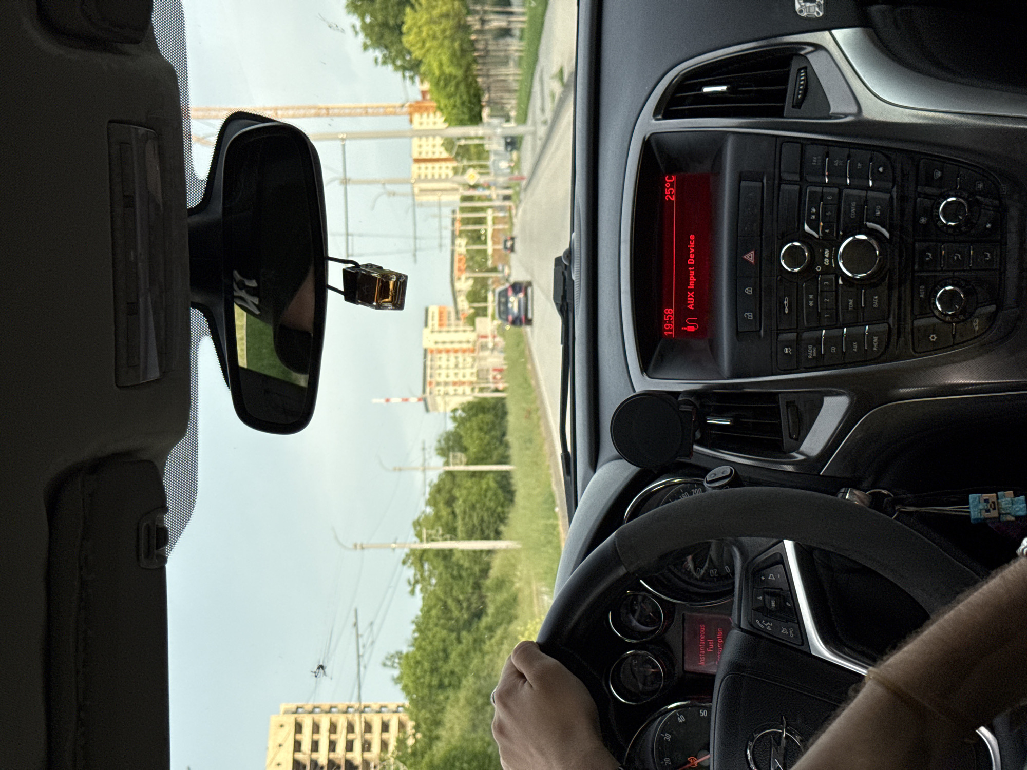 The image, taken from inside a car, captures a view of the road ahead in Pleven, Bulgaria. In the foreground, a driver is visible holding the steering wheel of a vehicle, identified as an Opel based on the steering wheel logo. The dashboard is prominent, displaying a car entertainment system set to AUX input at 19:58, with an ambient temperature of 25°C. The central console features numerous buttons and knobs.
The rearview mirror hangs from the windshield, slightly obscuring the view. Through the windshield, the road ahead is lined with trees, utility poles, and medium-rise apartment buildings. In the distance, a construction crane is visible. The image appears to be taken during daylight, under a clear sky. The specific location within Pleven is not discernible from the visual data.