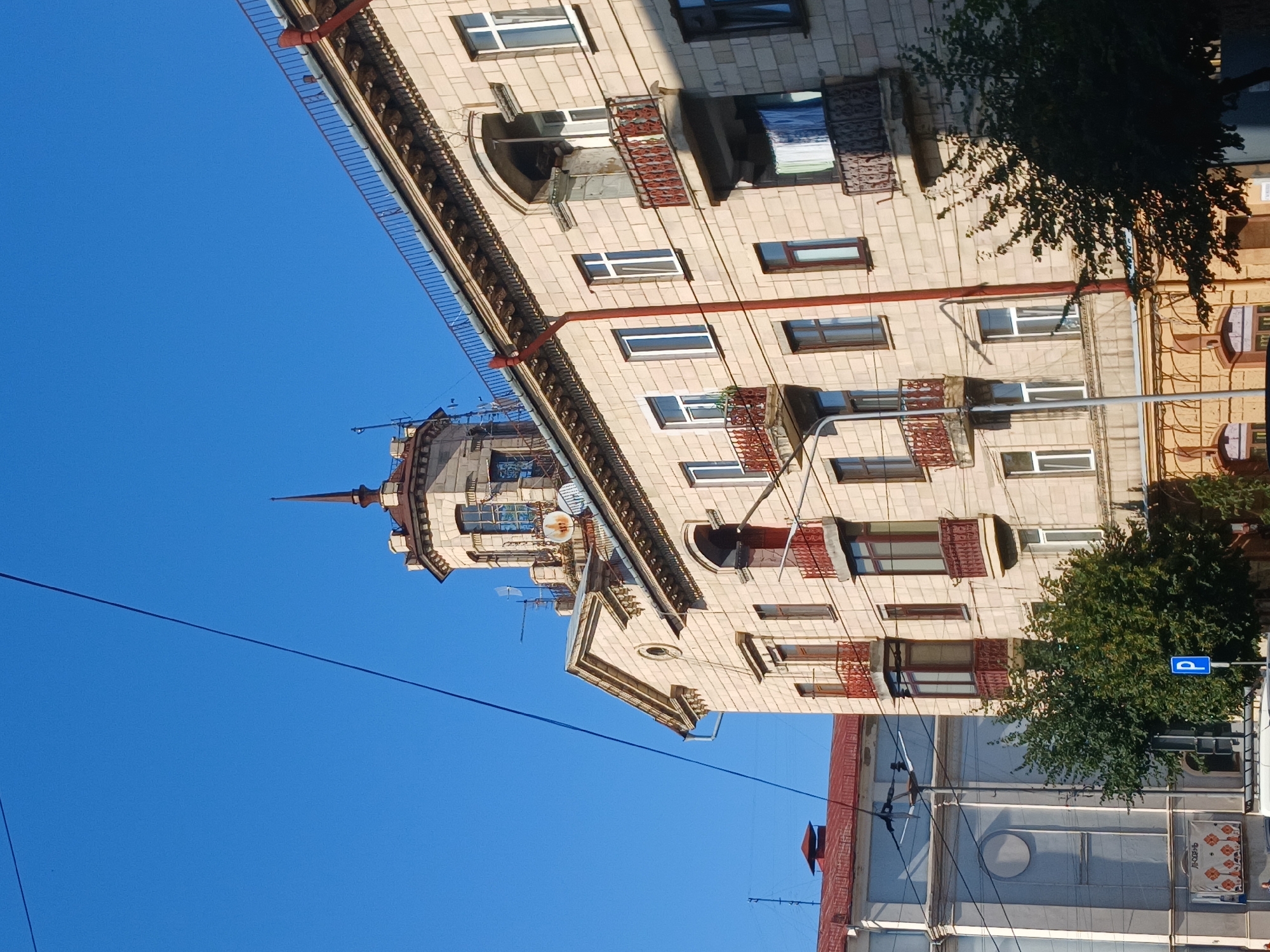 This is a slightly low-angle, exterior, daylight shot of an old, light-colored brick building in Chernivtsi, Ukraine. The building has a classical architectural style, with a prominent tower topped by a spire, several balconies with ornate railings, and numerous windows. A red drainpipe runs vertically down the facade of the building. Electrical wires are visible in the sky. In the foreground, to the left of the building, is another building with a red roof, and a street sign indicating parking. Trees with green foliage are also present, partially obscuring the lower levels of the buildings. The sky is a clear, bright blue.