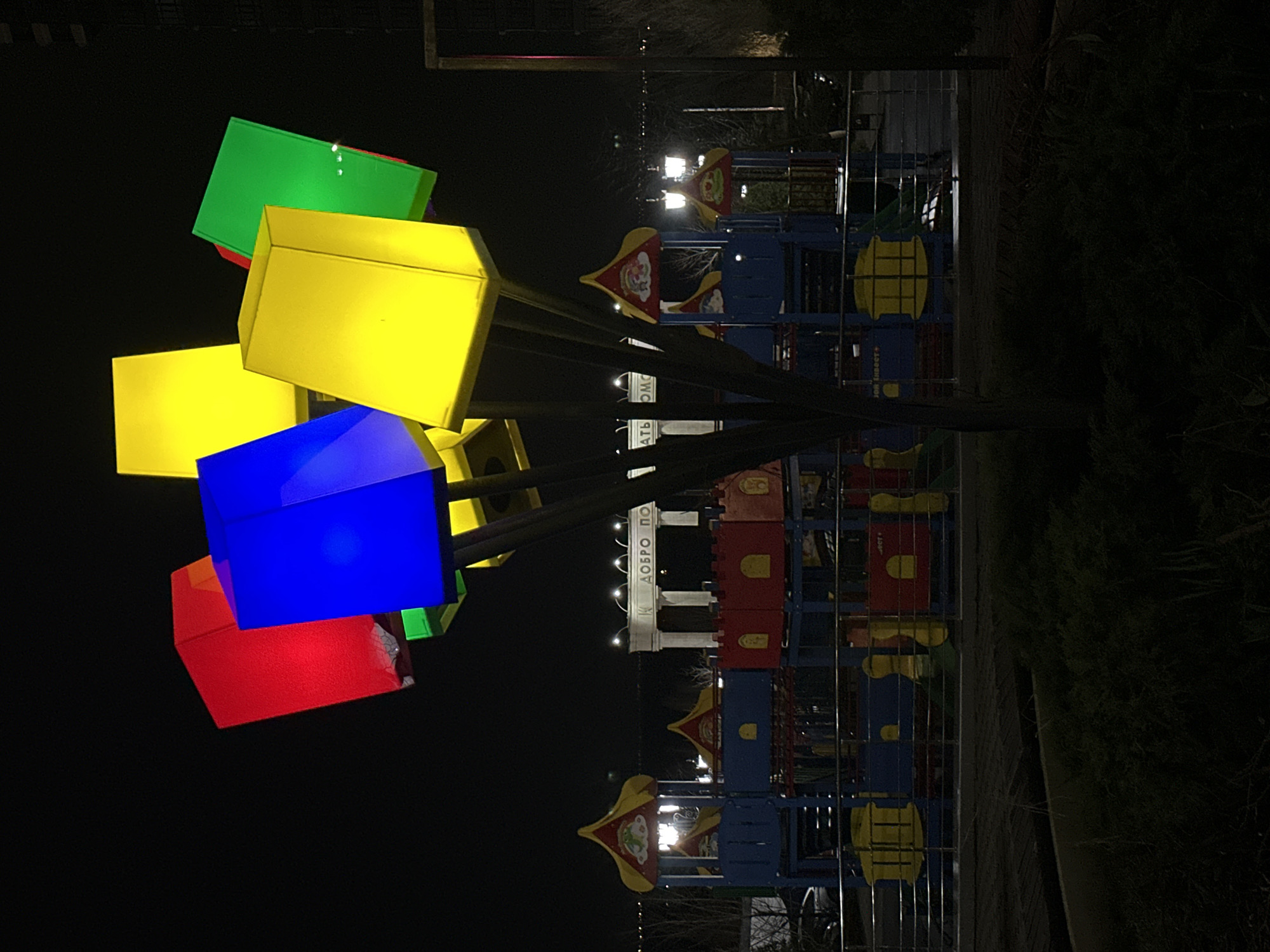 At night in Rostov-on-Don, Russia, a colorful playground is illuminated. A sculpture or light fixture composed of stacked, brightly colored cubes (red, blue, yellow, and green) dominates the left side of the frame. To the right, a metal-framed playground structure features red, yellow, and blue elements, with decorations. In the background, illuminated columns can be seen with writing, possibly "ДОБРО ПО" on top of them. Vegetation lines the right edge of the frame, and the rest of the background is dark.