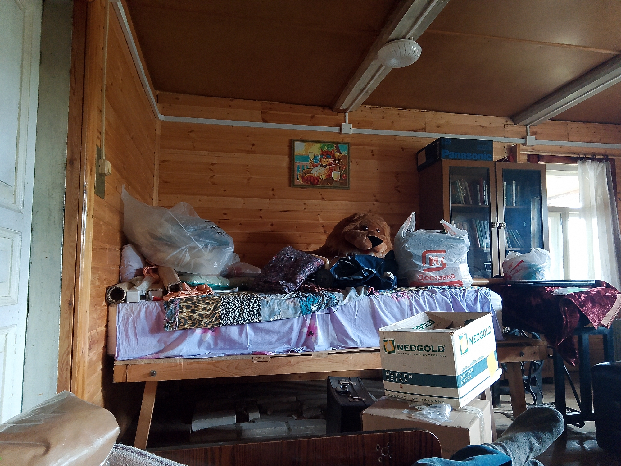 The image depicts a cluttered interior room, likely a bedroom or living space, with wooden paneled walls and ceiling. A bed is visible in the foreground, covered with a light purple sheet and various patterned blankets and items, including a leopard-print fabric. A large stuffed lion sits on the bed. Several plastic bags, some containing items, are scattered around the bed and on a nearby table. A bookshelf filled with books is to the right, topped with a Panasonic box. A framed picture of cartoon cats drinking is hanging on the wall. A window with sheer curtains is visible on the right side of the frame. The floor appears to be made of concrete or rough material with some scattered boxes and items. The overall impression is one of disarray and storage. The location is identified as the city of Kovrov, Russia.