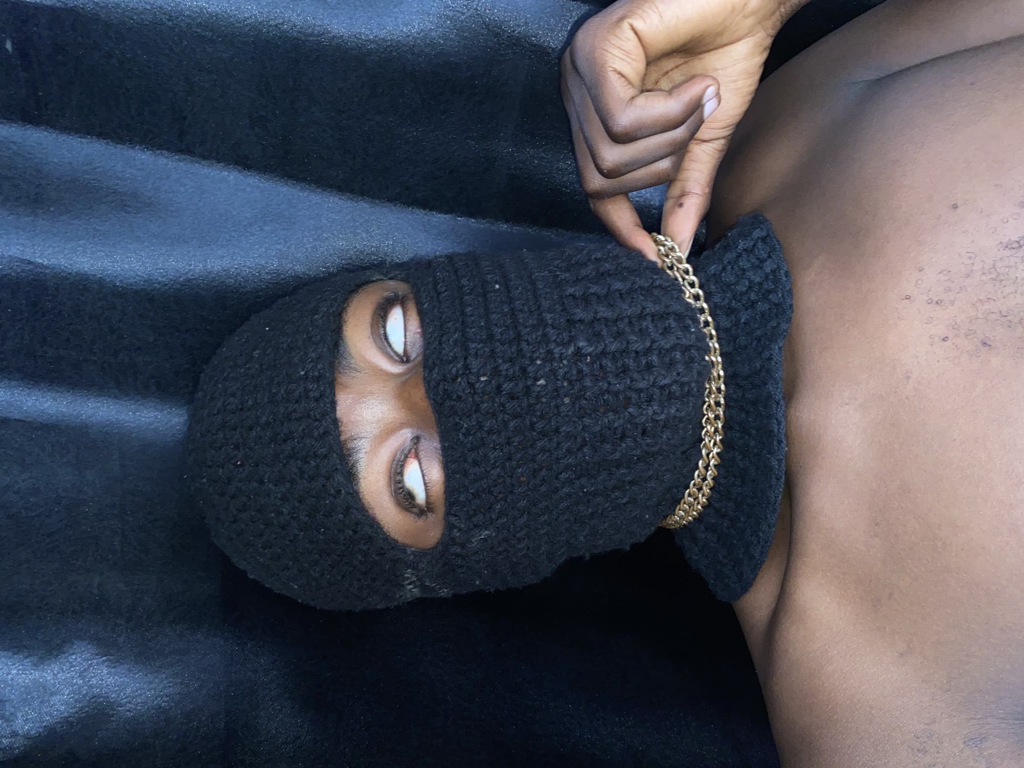 A person wearing a black knitted balaclava and gold chain lies on a dark surface that appears to be draped fabric. One hand is visible, holding the gold chain near the collar of the balaclava. The person's eyes are visible through the openings in the balaclava. The subject is partially shirtless. There is no explicit evidence to indicate this image was taken in Barakin Akawo, Nigeria.