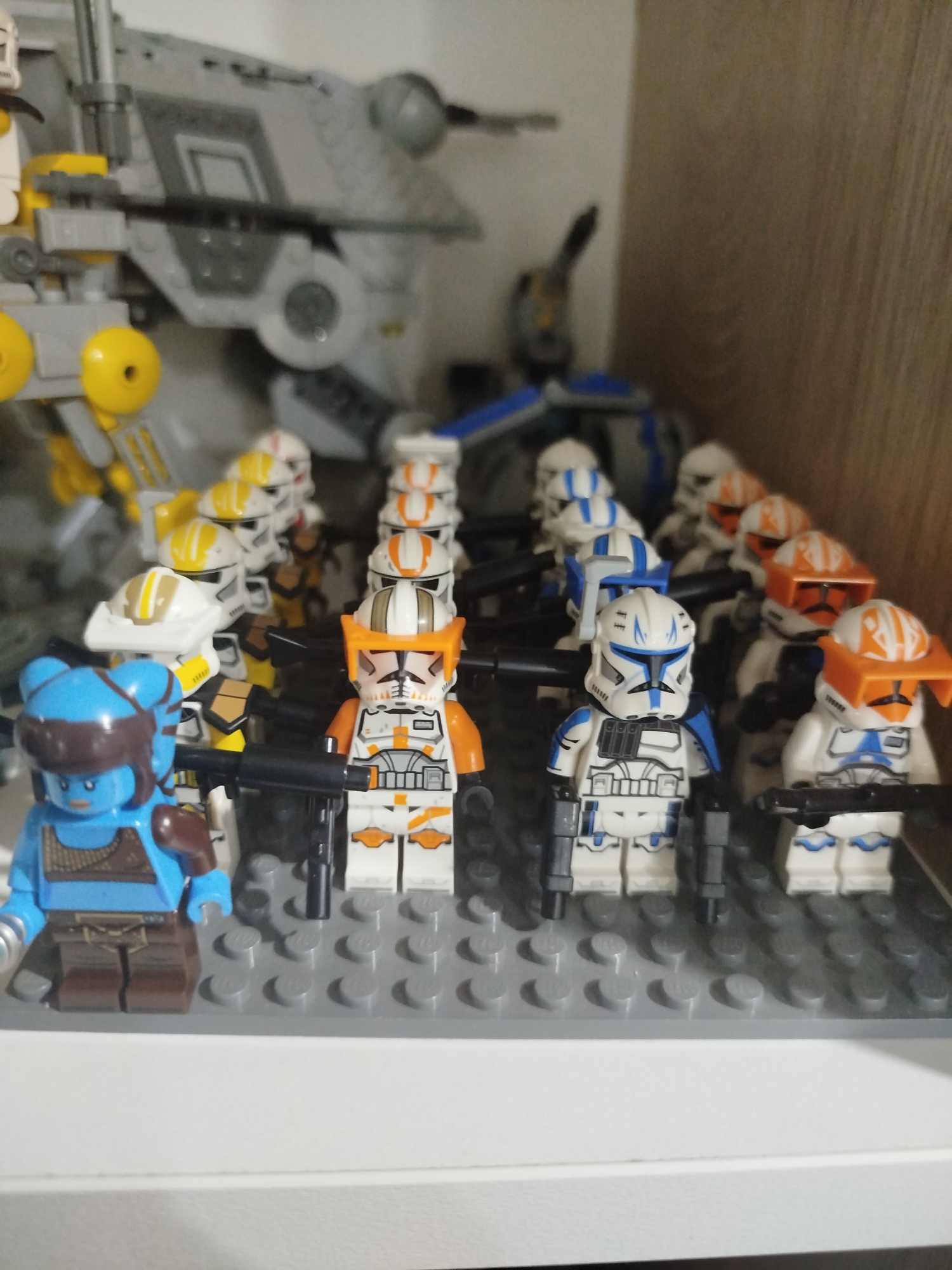 A collection of Lego Star Wars figures are displayed on a grey Lego base plate. In the foreground, a blue-skinned Lego figure with brown attire stands on the left. To its right, several Lego clone troopers in white and orange or white and blue armor are lined up, some holding black Lego blasters. In the background, a larger grey Lego vehicle, resembling an AT-AP walker, is partially visible. The figures and vehicle are placed on a shelf with a light brown wooden background. This image does not depict any location in Mirny, Russia.