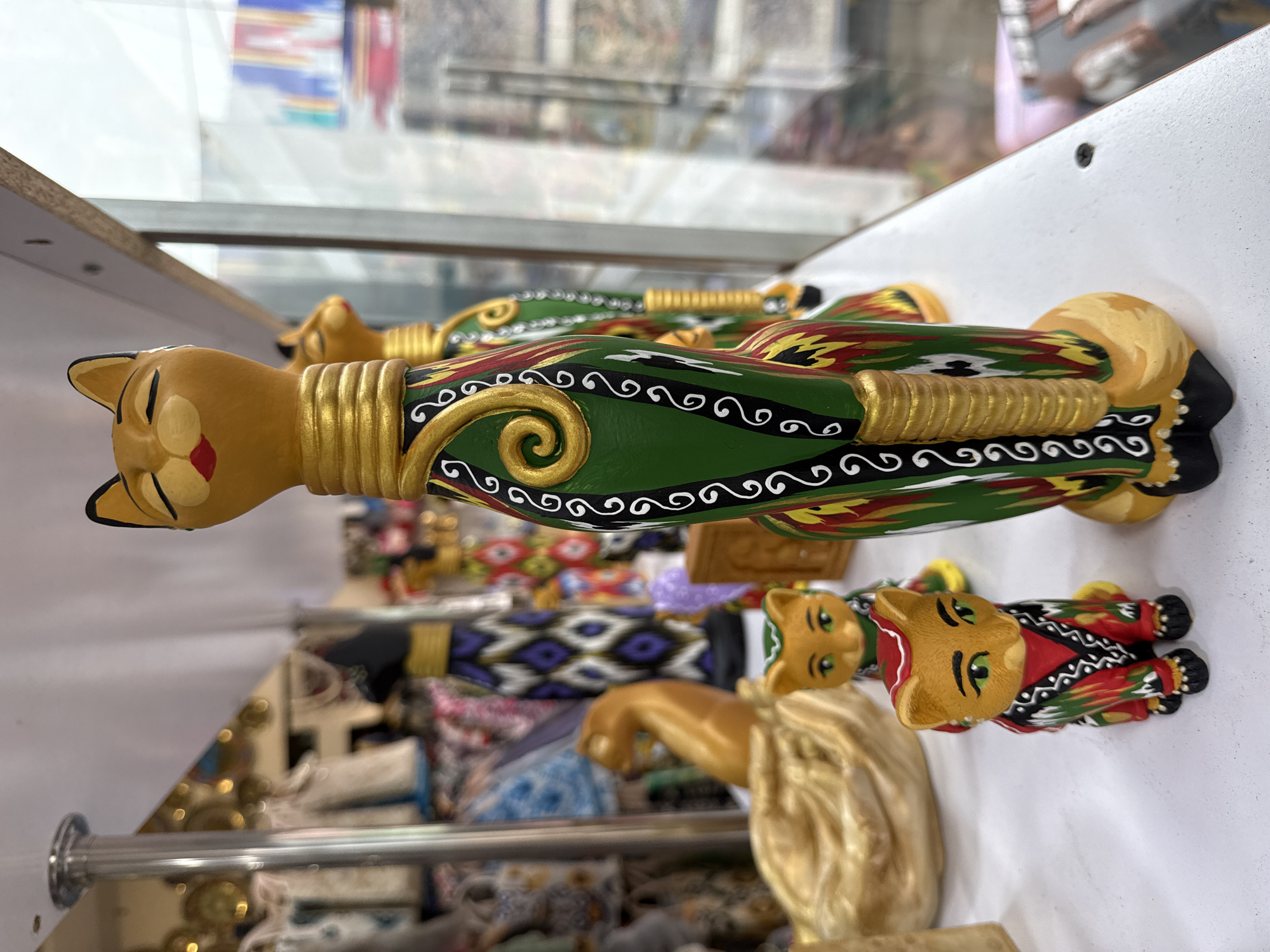 The image shows a row of intricately decorated wooden cat figurines displayed for sale, likely in a shop or market in Tashkent, Uzbekistan. The cats are lying on their sides on a white surface, potentially a shelf. Each cat features a yellow head and tail, with the body adorned in bright colors (green, red, and black) and swirling patterns. The decorations appear to be in a traditional Uzbek style. Some cats have gold accents on their necks. In the background, there's a glimpse of other merchandise, possibly textiles with colorful patterns. The presence of people and other items in the background suggests a bustling commercial environment. The image is taken at an angle, presenting a slightly blurred view.