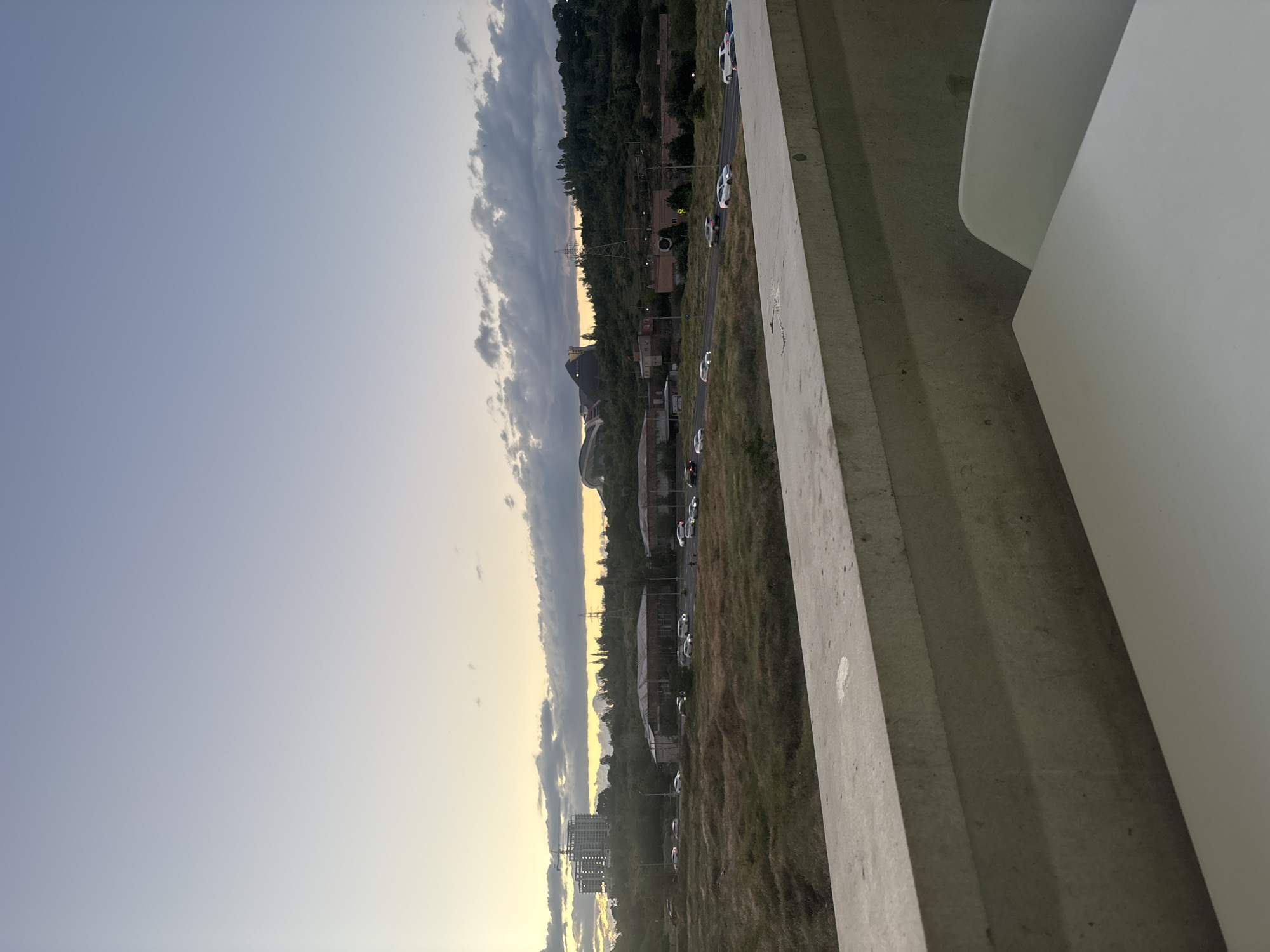 The image presents a wide landscape view captured from an elevated position, looking out over an area in Yerevan, Armenia, during what appears to be late afternoon or early evening. The sky dominates the upper portion, displaying a gradient from a warm, soft yellow at the horizon to a clear, pale blue overhead. Scattered clouds, some dark and voluminous, are visible, particularly along the horizon line.
In the mid-ground, a mix of natural and urban elements unfolds. There are hills or elevated terrain covered in green trees and dry, grassy patches. Numerous buildings, ranging from multi-story structures to smaller industrial or residential units, are interspersed within the landscape. A road or highway runs through the scene, with several white cars parked or driving along its side. A tall transmission tower or power line pylon is a notable feature in the distance.
The foreground on the right side of the image features a substantial concrete structure, likely a bridge railing or a building's parapet, indicating the viewpoint's height. Partially visible in the extreme right foreground is a curved, white object, possibly part of a chair or furniture. The overall scene suggests a blend of natural environment with urban development, under a serene, transitional sky. No discernible text is visible.