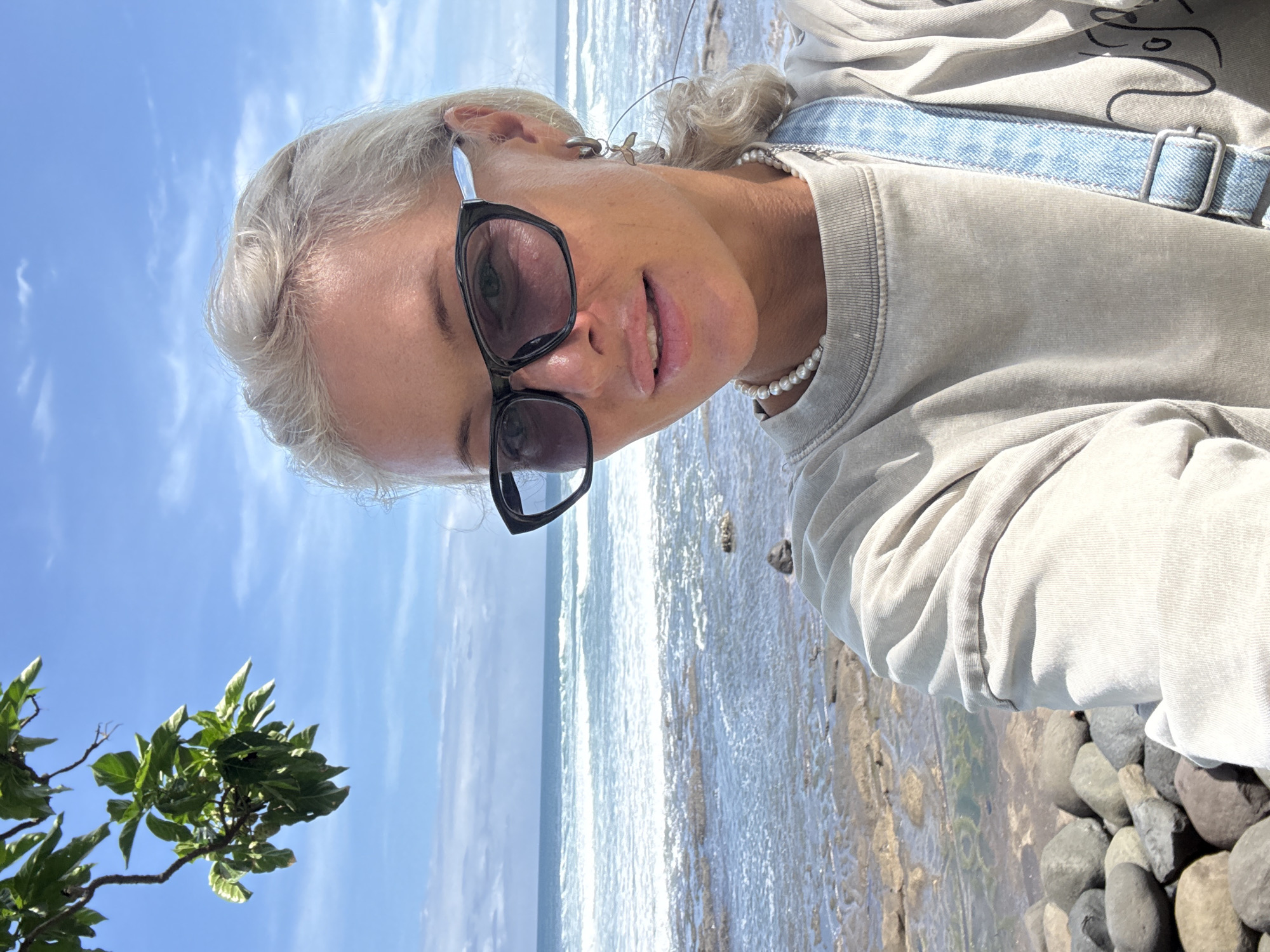 The image captures a woman at a beach in an unknown location. The woman is fair-skinned with blonde hair and is wearing sunglasses, a pearl necklace, and a light-colored shirt with a denim strap visible. She is positioned in the foreground, likely taking a selfie. The background shows the ocean with visible waves, a clear blue sky, and some green foliage to the left. To the right are a collection of pebbles and rocks along the shoreline.