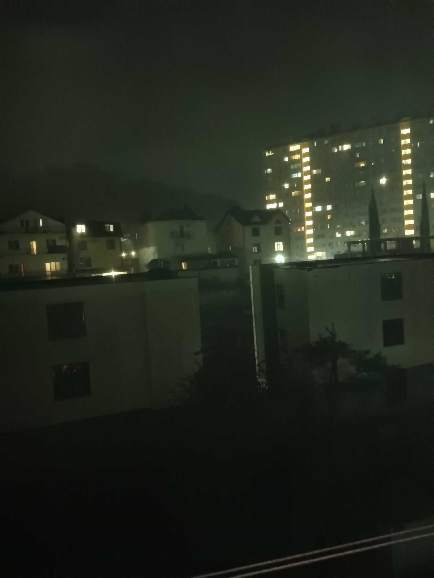 The image depicts an urban scene at night, likely in Sochi, Russia, characterized by a dark, overcast sky. Several buildings are visible, varying in height and architectural style. In the background, a tall, multi-story apartment building dominates the skyline, its windows illuminated with a warm, yellowish light, indicating occupancy. To the left of this prominent building, several smaller, lighter-colored residential structures are visible, some with lit windows as well. In the foreground and mid-ground, there are lower, more modern-looking white buildings, one of which has dark, rectangular windows. The overall impression is one of a residential area at night, with streetlights and internal building lights casting a glow in the darkness. The presence of what appear to be cypress trees adds a touch of natural element to the urban landscape. The image's low light conditions and slight blur suggest it may have been taken with a handheld camera at night. There are no people or specific actions depicted in the scene.