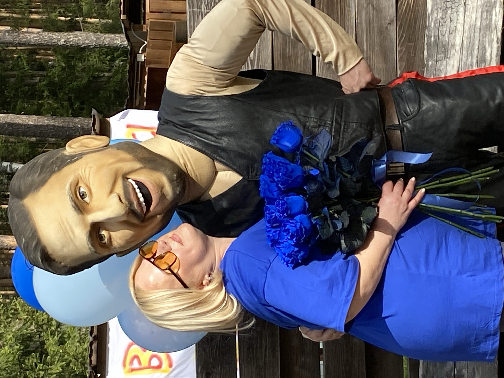 A photograph shows a woman holding a bouquet of blue roses next to a large, smiling figurine of a man. The woman has blonde hair, is wearing sunglasses and a blue t-shirt, and is standing on a wooden surface. The figurine is wearing a beige shirt, a black vest, and black pants with a red stripe down the side. There are also blue balloons in the shot. In the background, trees and a wooden structure can be seen, suggesting an outdoor location in Noviy, Russia.