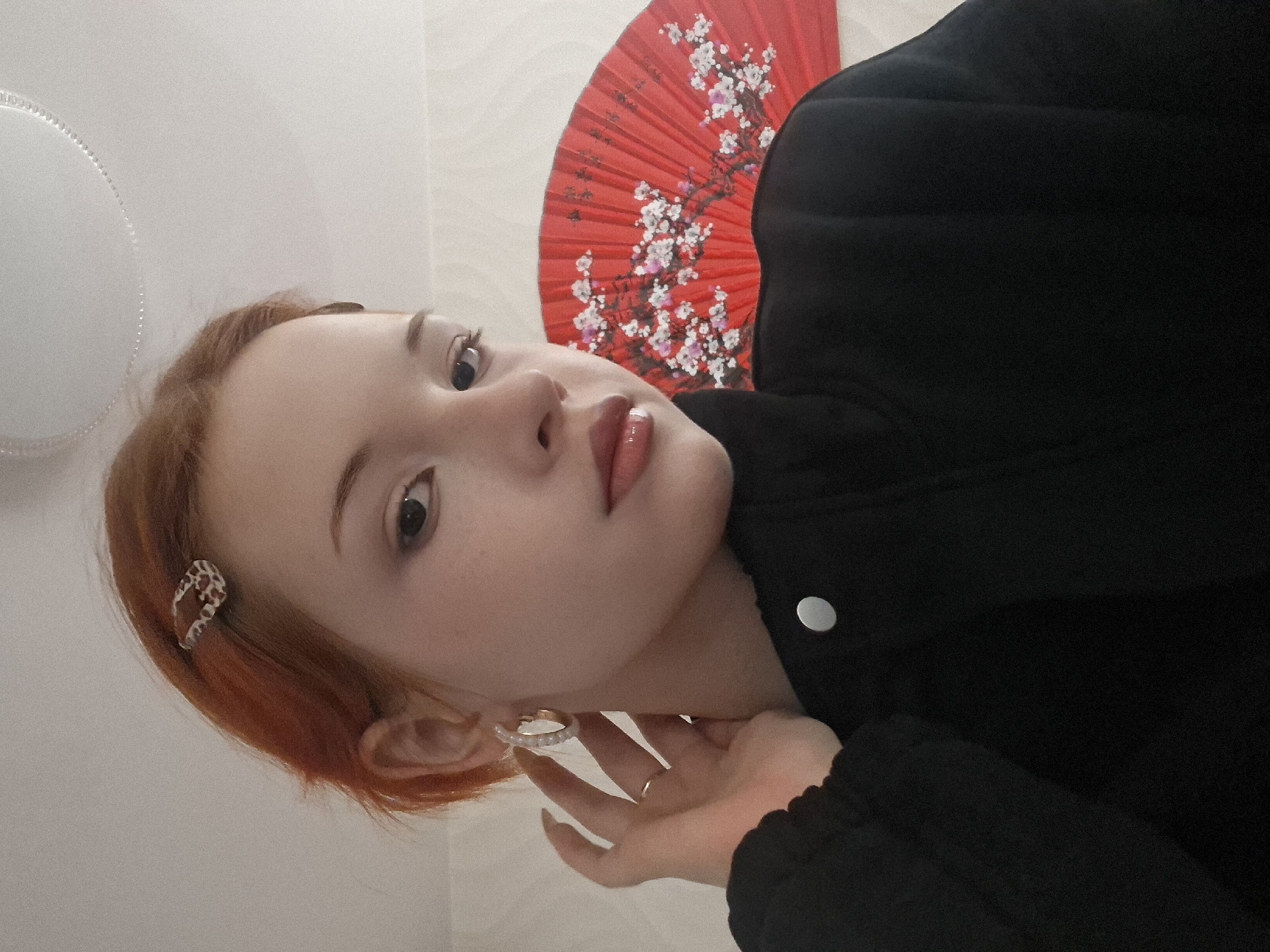 This image, taken in Chaikovsky, Russia, features a head-and-shoulders portrait of a young woman, captured with the camera rotated 90 degrees counter-clockwise. She has short, reddish-orange hair and fair skin, with light makeup accentuating her eyes and glossy lips. Her head is tilted, and she looks upwards and slightly to the left from the viewer's perspective, with a calm and neutral expression.
She is wearing a black jacket, which appears to be padded or textured. In her short hair, on the left side of her head (viewer's left), there is a patterned hair clip, possibly with an animal print. A decorative hoop earring with clear stones is visible on her right ear (viewer's left). Her right hand is positioned near her face, and a simple gold-colored ring is worn on her ring finger.
Behind her head and shoulder, a vibrant red folding fan with an intricate white and pink cherry blossom design is prominently displayed against a light-colored wall. To the far left of the frame, a partial view of a circular, white light fixture or lamp with a textured edge is visible. The scene is indoors, well-lit, and appears to be a casual portrait. There is no visible text in the image.