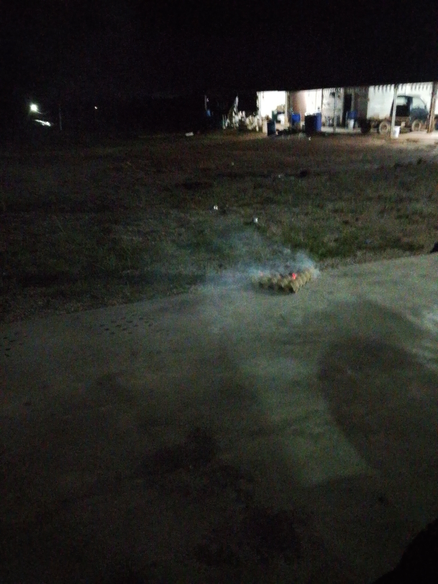 The image shows what appears to be burning material on a paved surface. The material is a small rectangle shape and is surrounded by smoke. The background is dark, but a lit-up structure with several objects and a truck can be seen in the distance. It is nighttime. The location is Bukit Ketok, Indonesia.