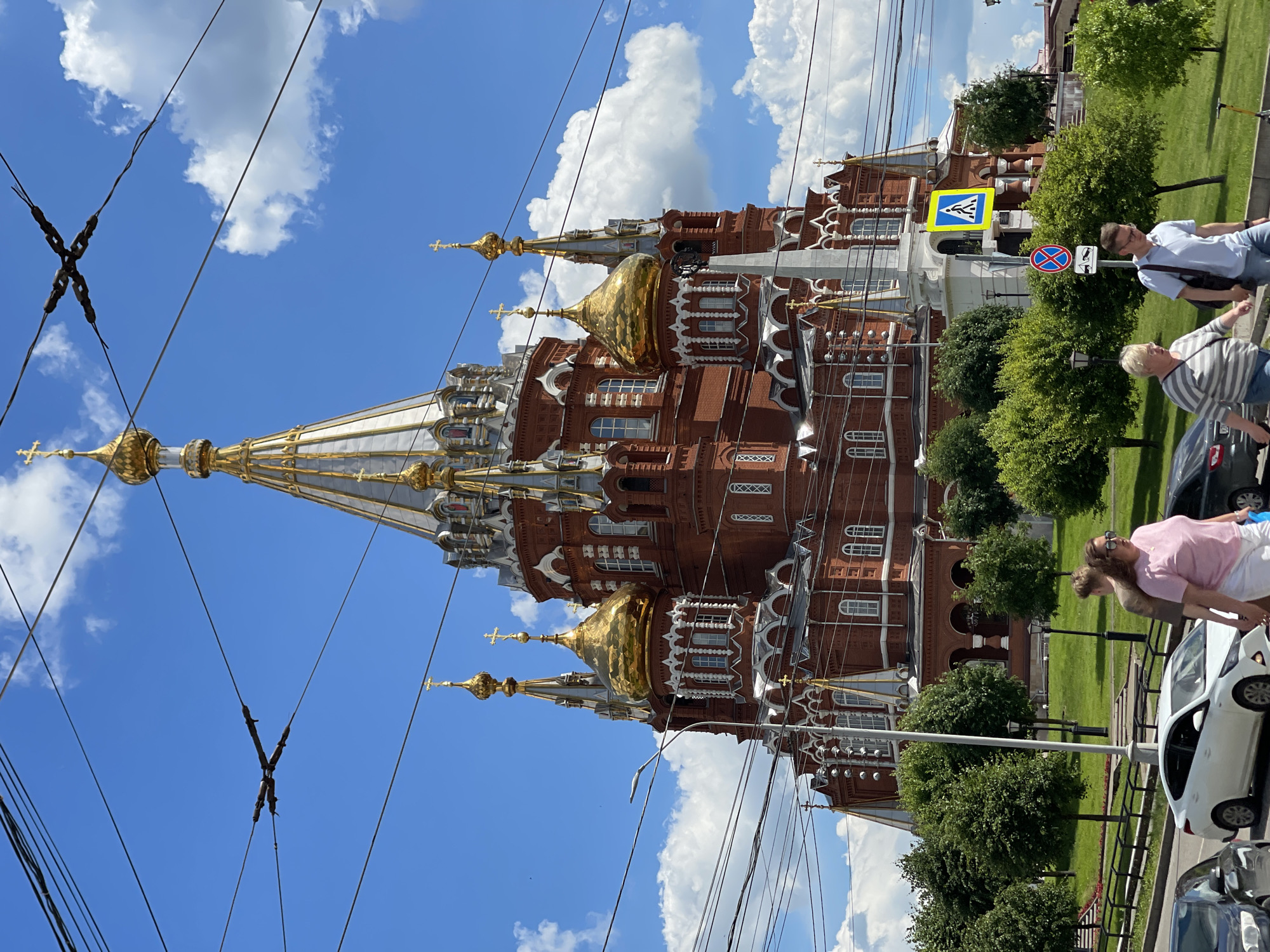 A large, red brick cathedral with gold domes and white architectural details stands against a bright blue sky with fluffy white clouds. Overhead, a complex network of black electrical wires crisscrosses the frame. In front of the cathedral, a green grassy lawn is dotted with trees. A sidewalk runs along the edge of the lawn, and several people are walking on it. A white car is parked on the street in the foreground. A pedestrian crossing sign is visible on the right side of the image. The overall scene suggests a sunny day in a city, with the cathedral being the prominent landmark.