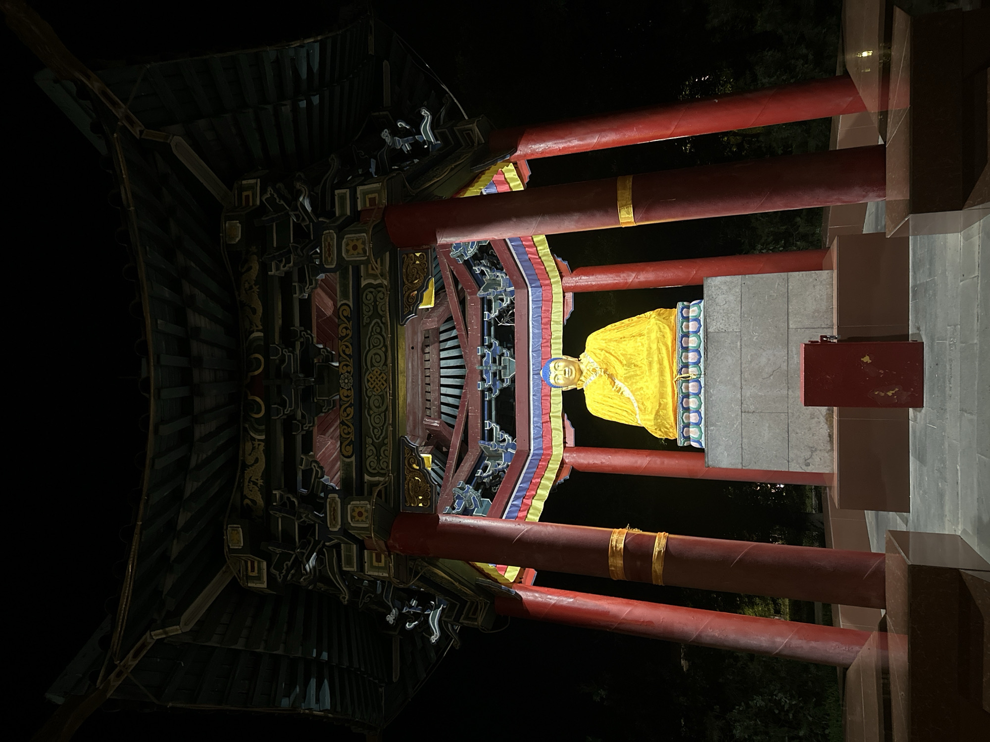 The image depicts a brightly lit Buddhist statue at night, housed within a colorful, traditional East Asian style pavilion. The statue, appearing to be of Buddha, is draped in a vibrant yellow cloth and has a blue headdress. It sits atop a tiered pedestal made of stone and patterned tiles. The pavilion itself is constructed with prominent red pillars and a dark, multi-layered roof adorned with intricate carvings and colorful decorative bands of yellow, blue, and red. The scene is set against a dark background, suggesting it is nighttime, with some foliage visible behind the structure. In the foreground, there are dark reddish-brown platforms and what appears to be a red metal box. This structure is located in Elista, Russia, a city known for its significant Buddhist population and numerous pagodas and stupas.