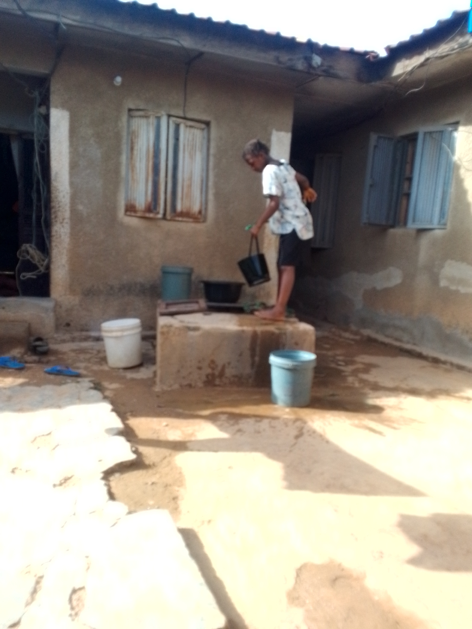 An outdoor daytime scene shows a person standing on a raised concrete structure in front of a weathered building, possibly in the city of Unknown, Unknown. The person is pouring water from a black bucket into a larger black container on the structure. Several other buckets are scattered around the base. The building has two windows with metal coverings or shutters. A pair of sandals rests near the building entrance. The ground appears wet.