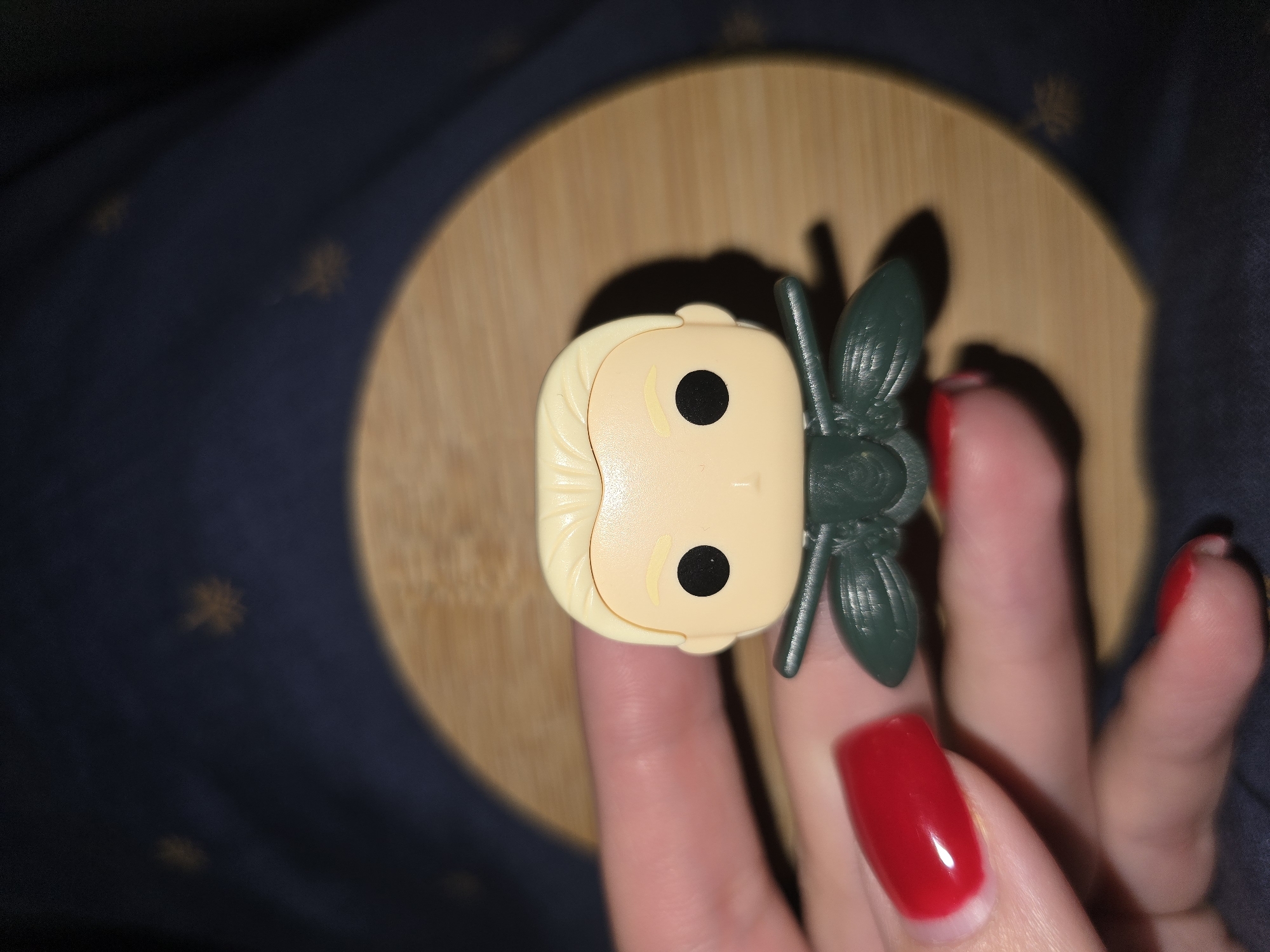 A close-up, top-down shot shows the head of a Funko Pop collectible figure held between two fingers of a person's hand. The figure has blonde hair and black eyes, with a green accessory resembling wings or antennae attached to the back of its head. The hand has bright red nail polish on its fingernails, with one finger visible beneath the figure and another finger to the right. The figure and hand are positioned on a round, light-brown wooden surface, which is itself on top of a dark blue fabric with a subtle pattern. The lighting suggests it may be indoors, with soft shadows cast by the figure and the wooden surface. The image appears to be a casual, personal photograph, possibly taken to showcase the collectible figure. There is no visible text in the image. The location context of Petropavlivska Borshchahivka, Ukraine, is provided externally and not directly evident from the visual cues in the photograph itself.