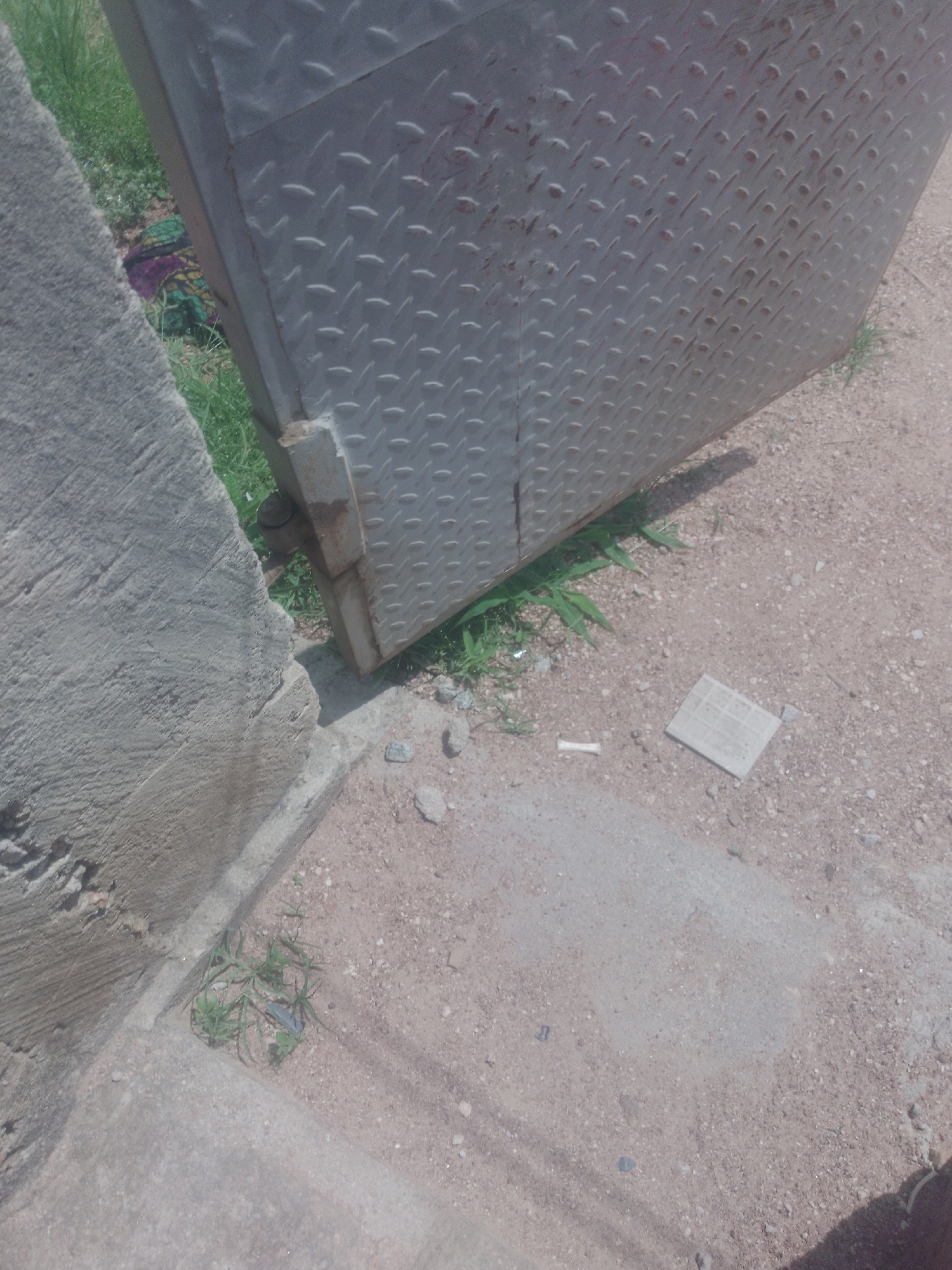 The image depicts an outdoor scene in Jauro Yinu, Nigeria. A large, metal diamond plate door or panel is partially open and rests on the ground. Adjacent to the metal structure is a rough, concrete wall or pillar. The ground is a mix of sandy soil, small stones, and patches of dry grass. A piece of folded paper or thin cardboard lies on the ground near the metal panel. There are no people visible in the image.