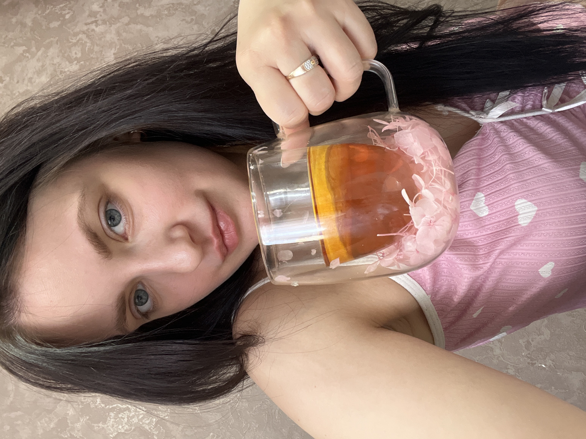 A woman with long, dark hair and light-colored eyes is holding a glass mug filled with a light brown liquid, possibly tea. The mug has a handle and decorative pink flowers. She is wearing a pink top with a white trim and a pattern of white hearts. A ring is visible on her ring finger. The background appears to be a light-colored wall. The location is Марковский, Russia.