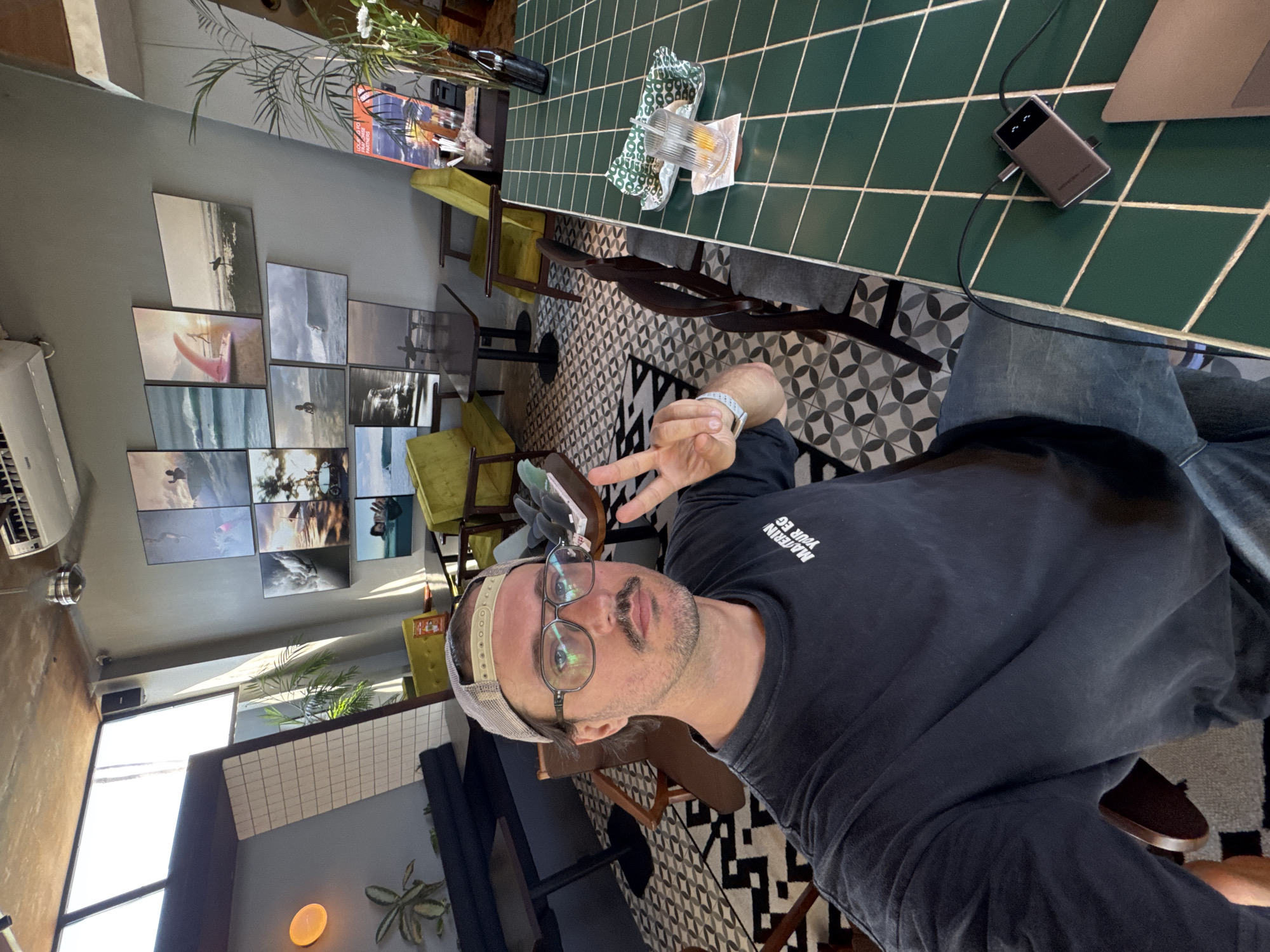 A man wearing eyeglasses and a baseball cap is sitting at a table in a building with various chairs, plants, and wall decorations. The table has a green tiled surface, on which are a glass of liquid, and a laptop docking station connected to a laptop. The floor has a black and white geometric pattern. The location is in Kuta Selatan, Indonesia.