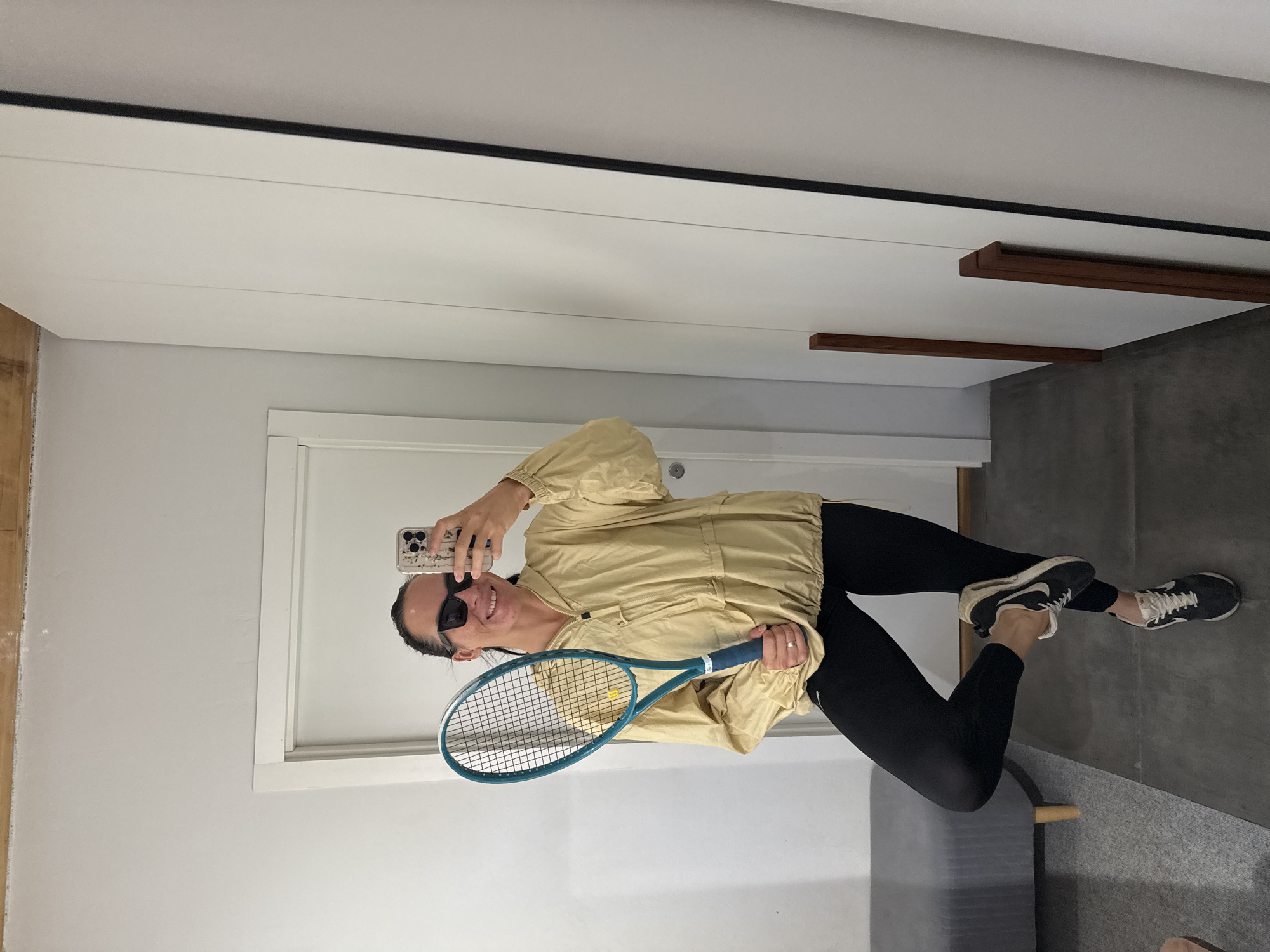 A woman is taking a selfie in a room that appears to be a home gym or dressing room. She is standing in front of a mirror and holding a tennis racket. She is wearing a light yellow hooded jacket, black leggings, and black and white athletic shoes. She has dark hair pulled back and is wearing dark sunglasses. The room has white walls and a white door. To the right of the woman is a large white mirrored closet door. To the left of the woman is a grey bench with wooden legs. The floor is covered with a dark grey mat. The woman is on the left side of the image, with her body angled towards the mirror. She is holding the tennis racket in her right hand, with the head of the racket above her shoulder. Her left leg is bent at the knee and her left foot is resting on her right ankle. The lighting in the room is bright and even. There is no indication of the specific location within the city of Chaikovsky, Russia.