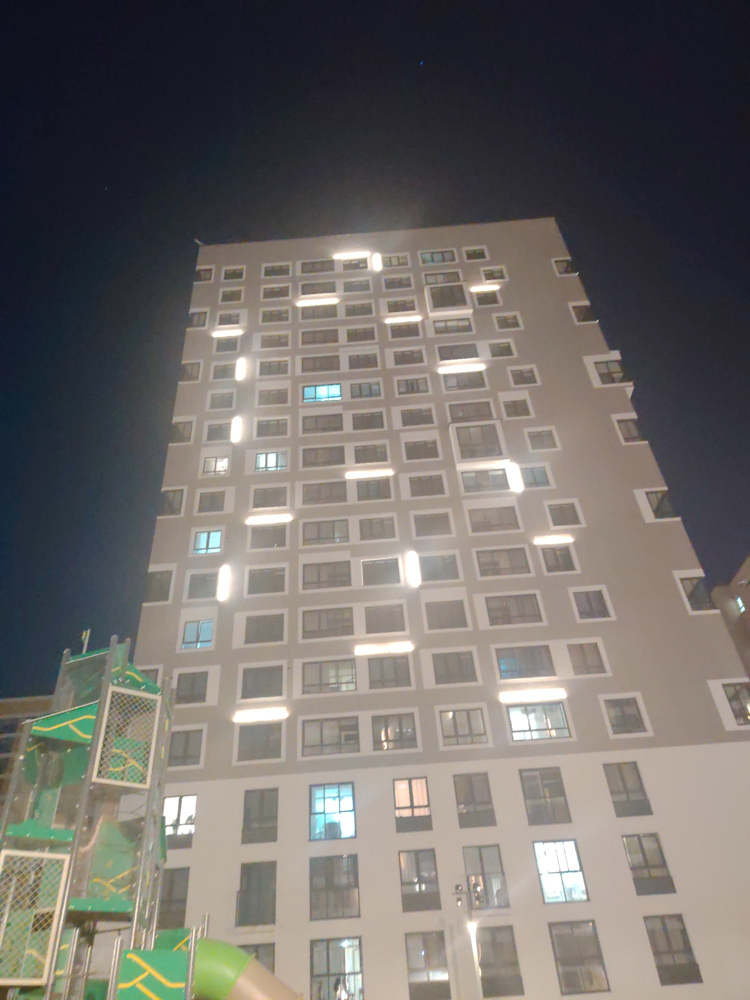 The image shows a high-rise apartment building at night. The building is light gray with a pattern of windows, some of which are illuminated. In the foreground, there is a children's playground with green and yellow structures. The sky is dark blue, indicating nighttime. The building's architecture is modern, with a staggered pattern of windows. There are no people visible in the image. This building is located in the city of Yekaterinburg, Russia, specifically in the Novokoltsovsky district, near the Ural Federal University.