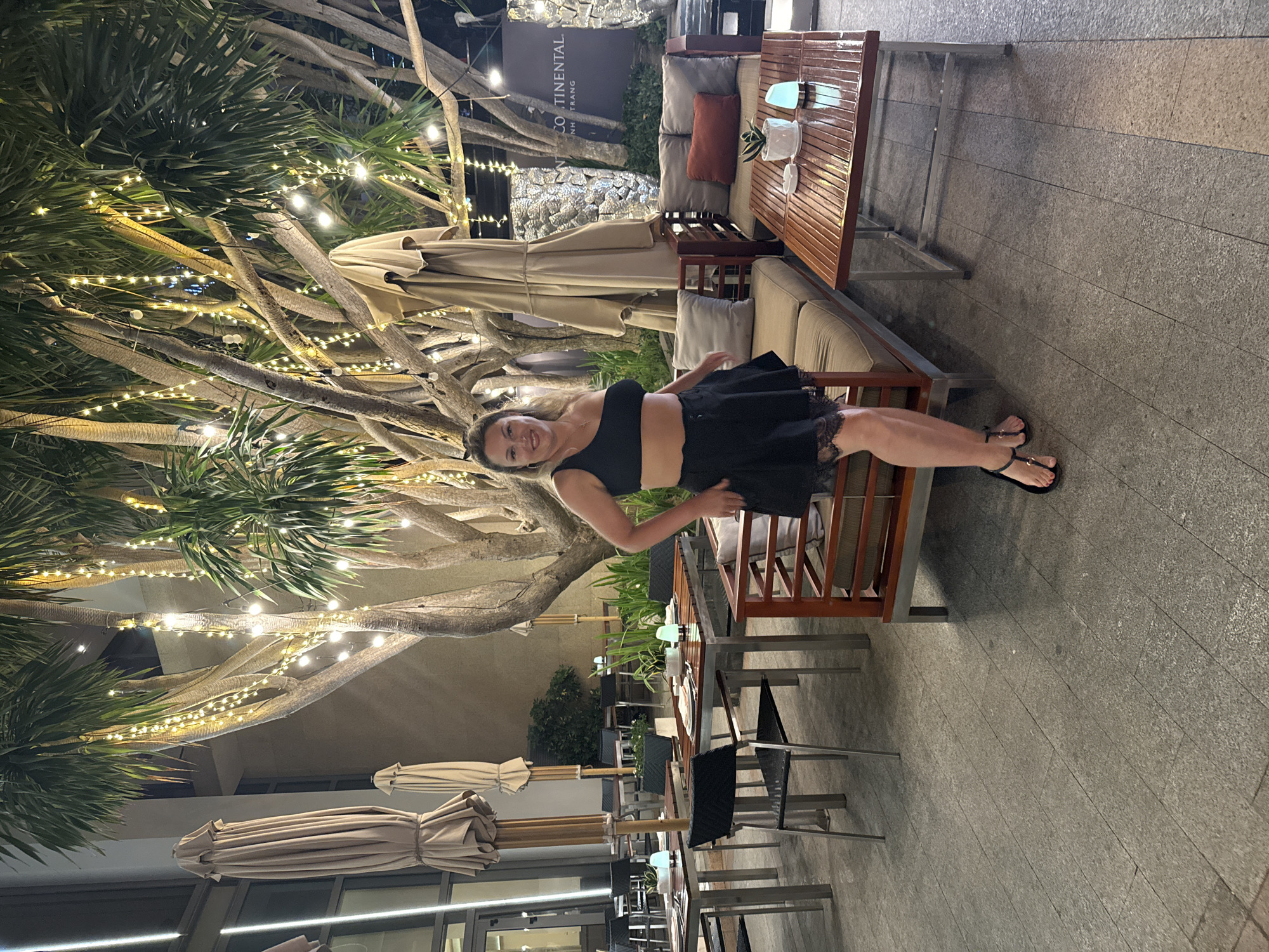An outdoor dining area in Nha Trang Ward, Vietnam, features a woman posing in a black top and skirt, positioned between a cushioned bench and a table. Overhead, a large tree is adorned with string lights. In the background, partially furled umbrellas line the perimeter. Furniture includes metal-framed chairs and tables. A sign reading "Continental Nha Trang" is visible in the backdrop. The ground consists of horizontally laid grey stone tiles.