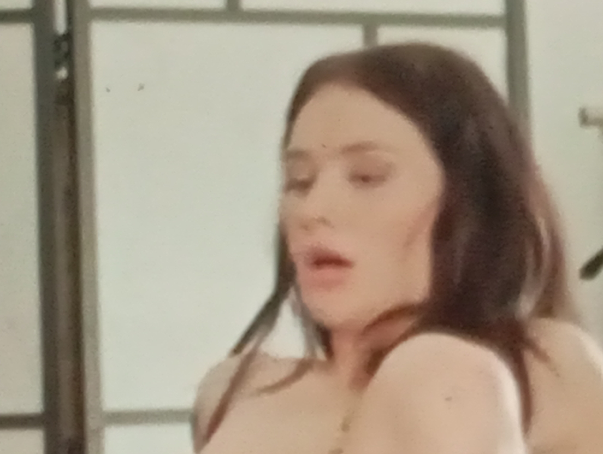 The image is a close-up of a woman's face and upper torso. She has long, dark brown hair, and her eyes are closed. Her mouth is slightly open, and she appears to be exhaling or speaking. The background is out of focus, but it appears to be a room with a light-colored wall and a vertical structure, possibly a screen or partition. There are no visible signs of the city of Sofiivska Borshchahivka, Ukraine, in the image.