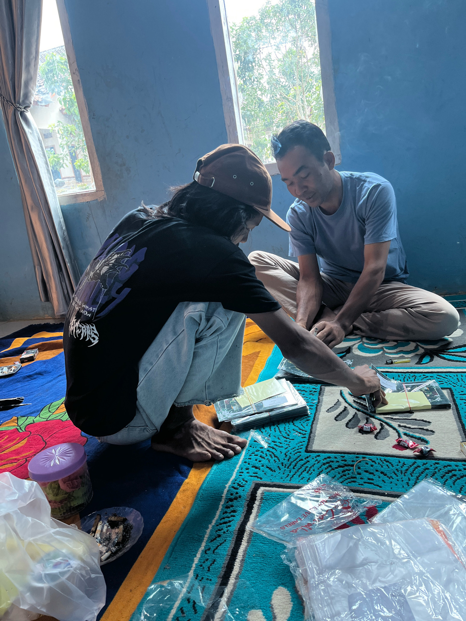 Two individuals are seated on a colorful rug inside a room with blue walls. One person, wearing a brown baseball cap and a black t-shirt with a graphic print, is crouched on the floor. The other person, wearing a light blue t-shirt and beige pants, is sitting cross-legged. They are both engaged in handling stacks of what appear to be cards or small booklets, some of which are wrapped in plastic. Scattered around them are various items, including small candies, a purple container with what looks like artificial flowers, a small dish with cigarette butts, and several plastic bags, some containing white items. The room has a window with a gray curtain on the left, and natural light streams in from the window, illuminating the scene. The overall impression is one of a casual activity taking place indoors. The image is set in Kebumen, Indonesia.