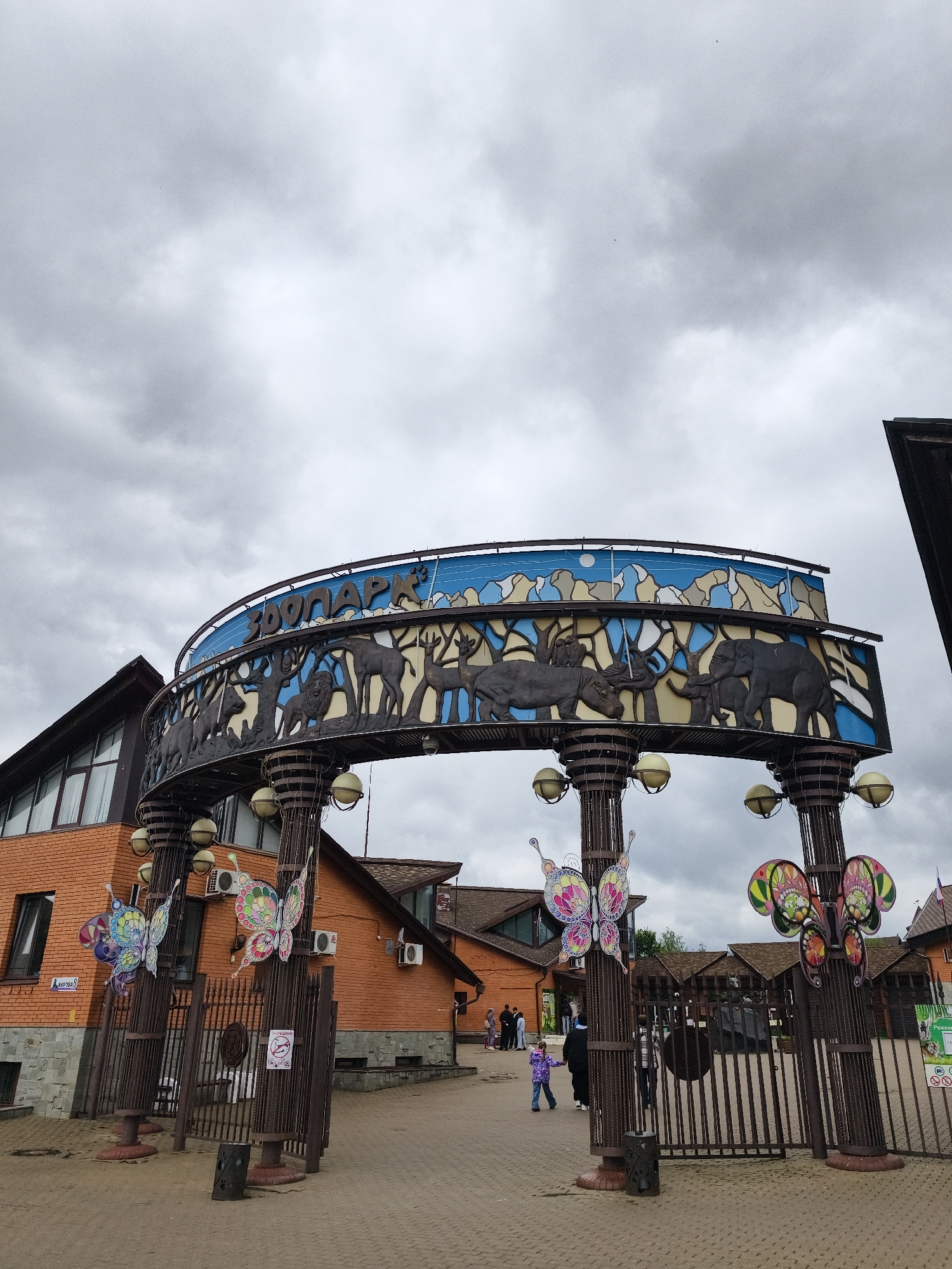 The image captures the entrance to a zoo or animal park on an overcast day in Izhevsk, Russia. A prominent, semi-circular archway spans the width of the entrance, serving as the main sign. This archway is decorated with a dark metal frieze depicting various animals in relief, including lions, deer/elk, hippopotamuses, rhinoceroses, and elephants, set against a stylized landscape of trees and hills. Above this, the word "ЗООПАРК" (ZOOPARK) is displayed in blue Cyrillic letters against a bright blue background with white cloud and mountain silhouettes.
The entrance features sturdy metal gates, currently open, flanked by tall, dark metal pillars. These pillars are intricately designed with a twisted metal pattern and are adorned with decorative, colorful butterfly cutouts that are illuminated by spherical light fixtures. Beyond the gates, several brick buildings with sloped brown roofs and large windows are visible, typical of an administrative or visitor center structure. Some air conditioning units are mounted on the exterior walls of these buildings.
A few people are present on the paved pathway leading into the park, including adults and a young child in a purple jacket walking towards the entrance. The ground is covered with interlocking pavers. The sky is heavily overcast with a uniform layer of grey clouds, indicating a cloudy daytime setting.
Visible text includes:
- "ЗООПАРК" (ZOOPARK) on the archway sign.
- "КИРОВА 8" (KIROVA 8), likely a street address, on a red sign on the brick building to the left.
- A white sign on the gate with a red circle and diagonal line over a burning cigarette, indicating "КУРЕНИЕ ЗАПРЕЩЕНО" (SMOKING PROHIBITED), and another symbol below it suggesting "NO LITTERING."
- A partially visible green sign on the right mentions "Режим раб..." (Operating hours...).
- A small white sign at the base of the right pillar states "ПОД ВИДЕОНАБЛЮДЕНИЕМ" (UNDER VIDEO SURVEILLANCE).